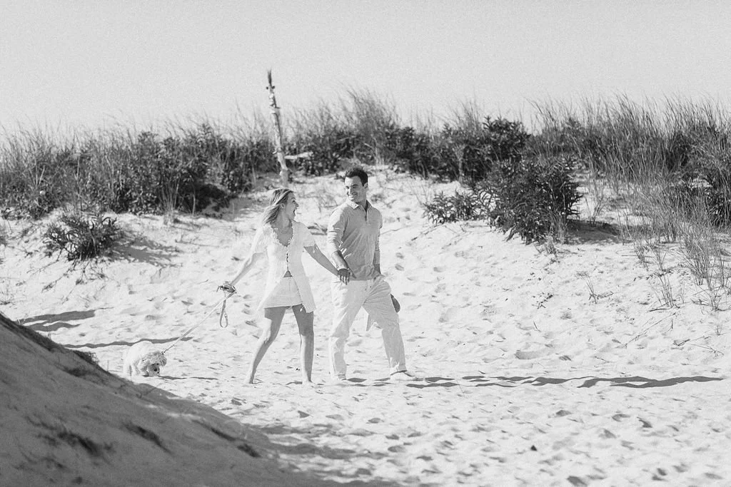 A couple walking along the beach, holding hands, with a small dog on a leash, dunes, and grasses in the background.