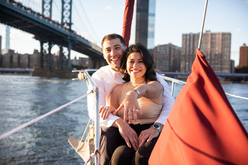 A smiling couple on a boat in New York City with a bridge and city buildings in the background during sunset.