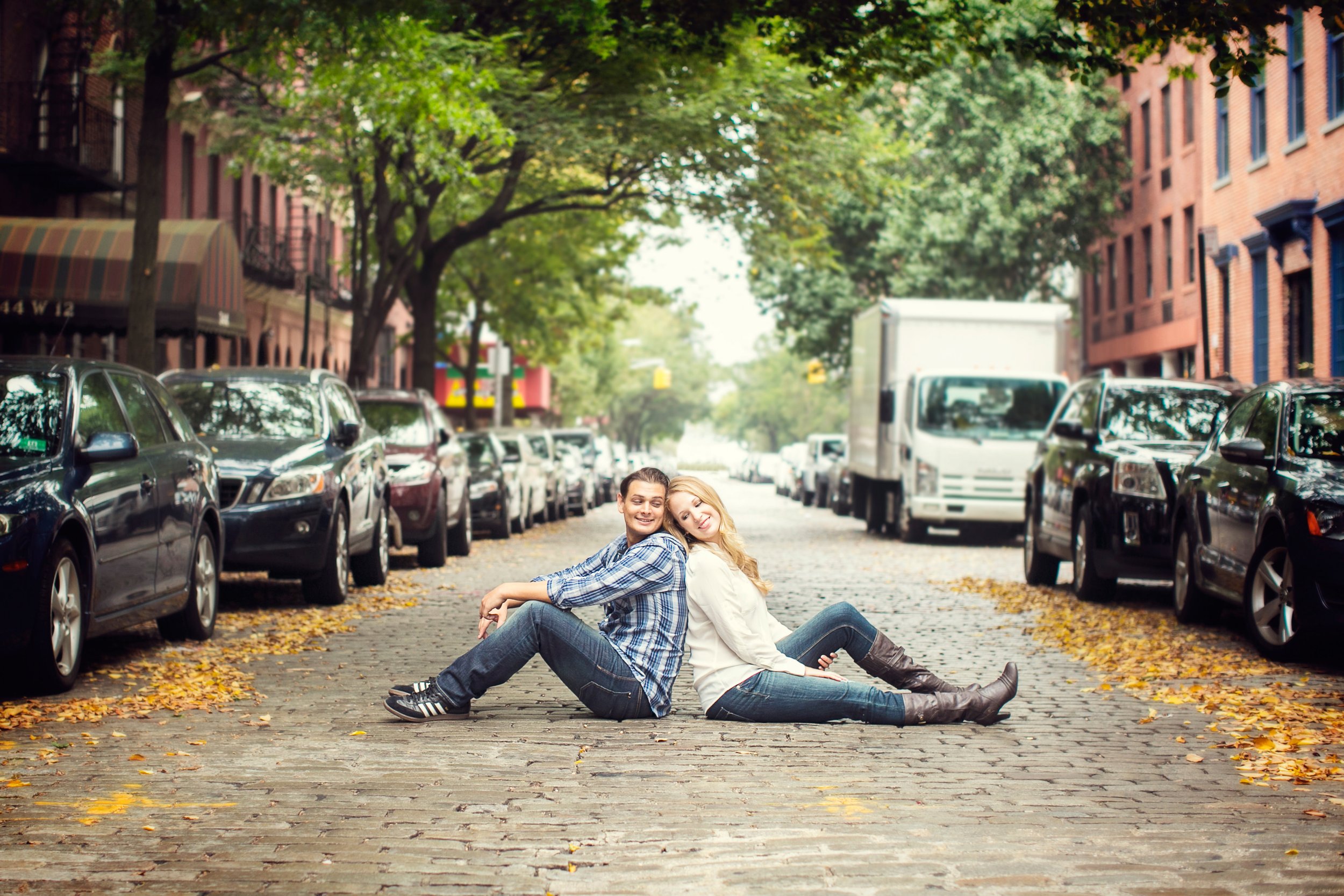 A couple sitting back-to-back on a cobblestone street lined with parked cars and trees in an urban neighborhood.