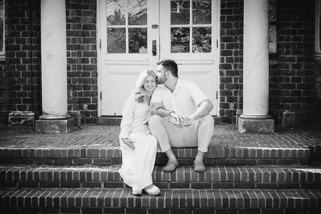 A smiling couple sitting on brick steps in front of a building with a door and window, embracing and sharing a tender moment.