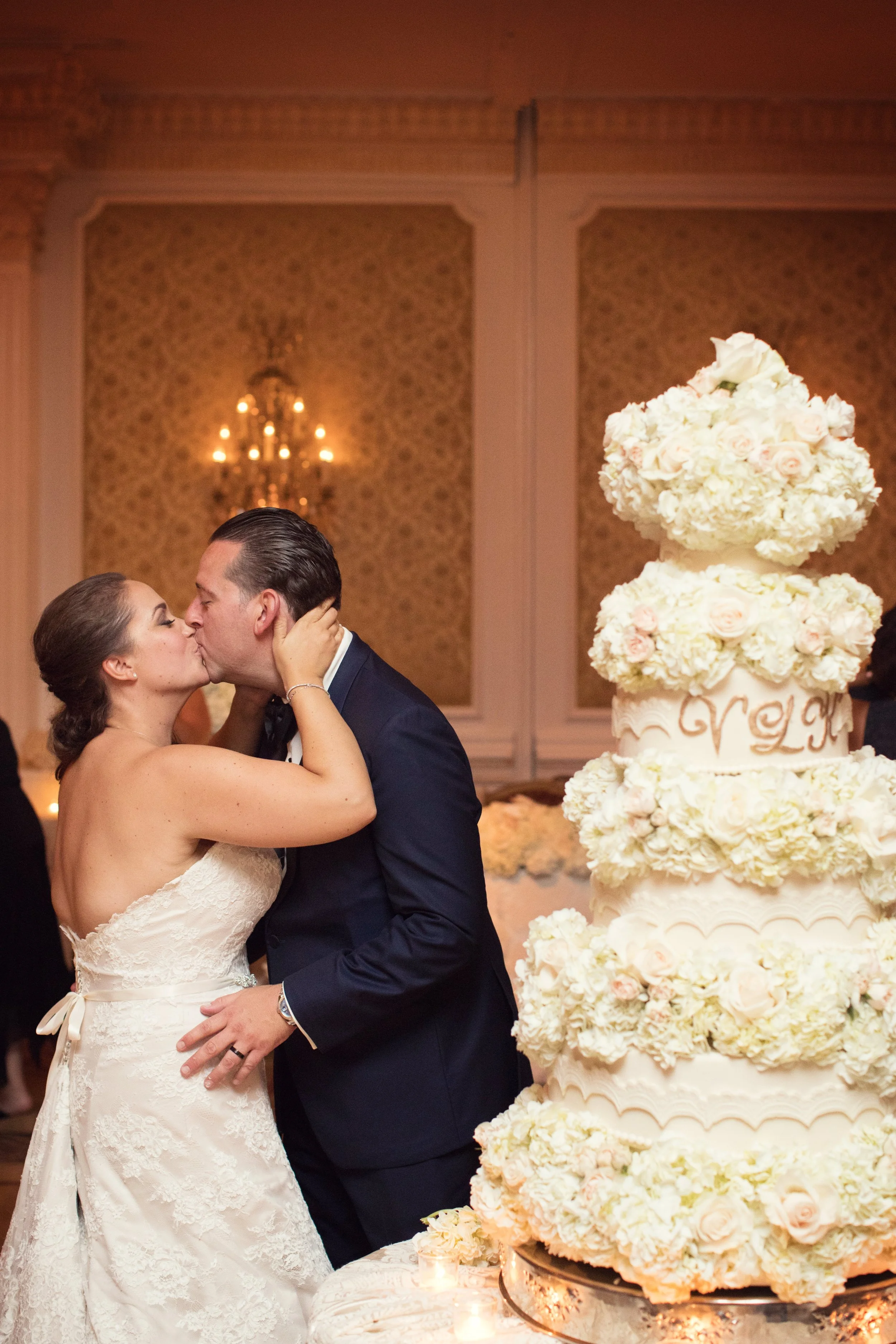 A bride and groom kiss in front of a large wedding cake decorated with white and pale pink flowers.