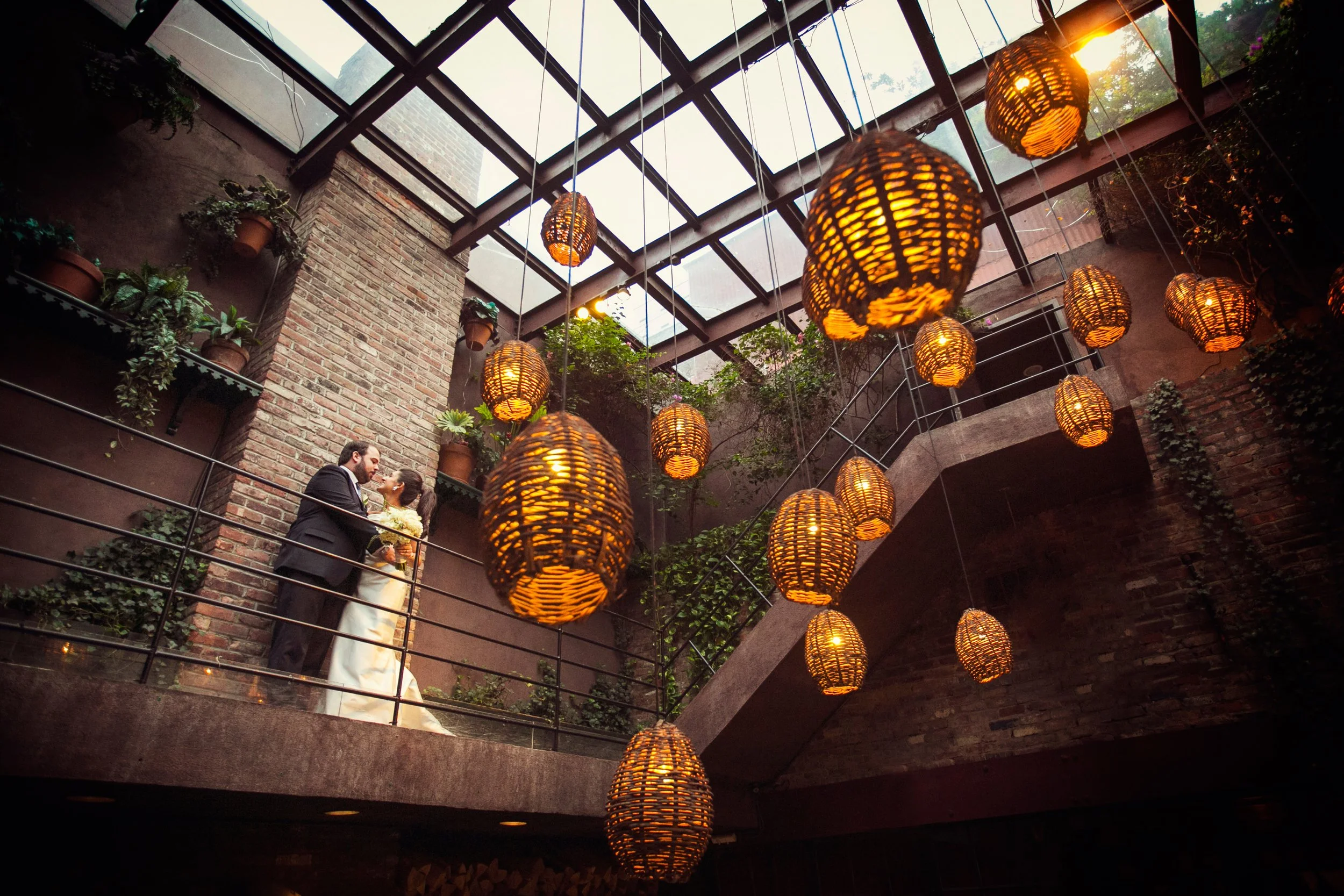 A bride and groom sharing a kiss on an indoor balcony with warm hanging wicker lanterns and greenery, in a rustic industrial venue with brick walls and a glass roof.