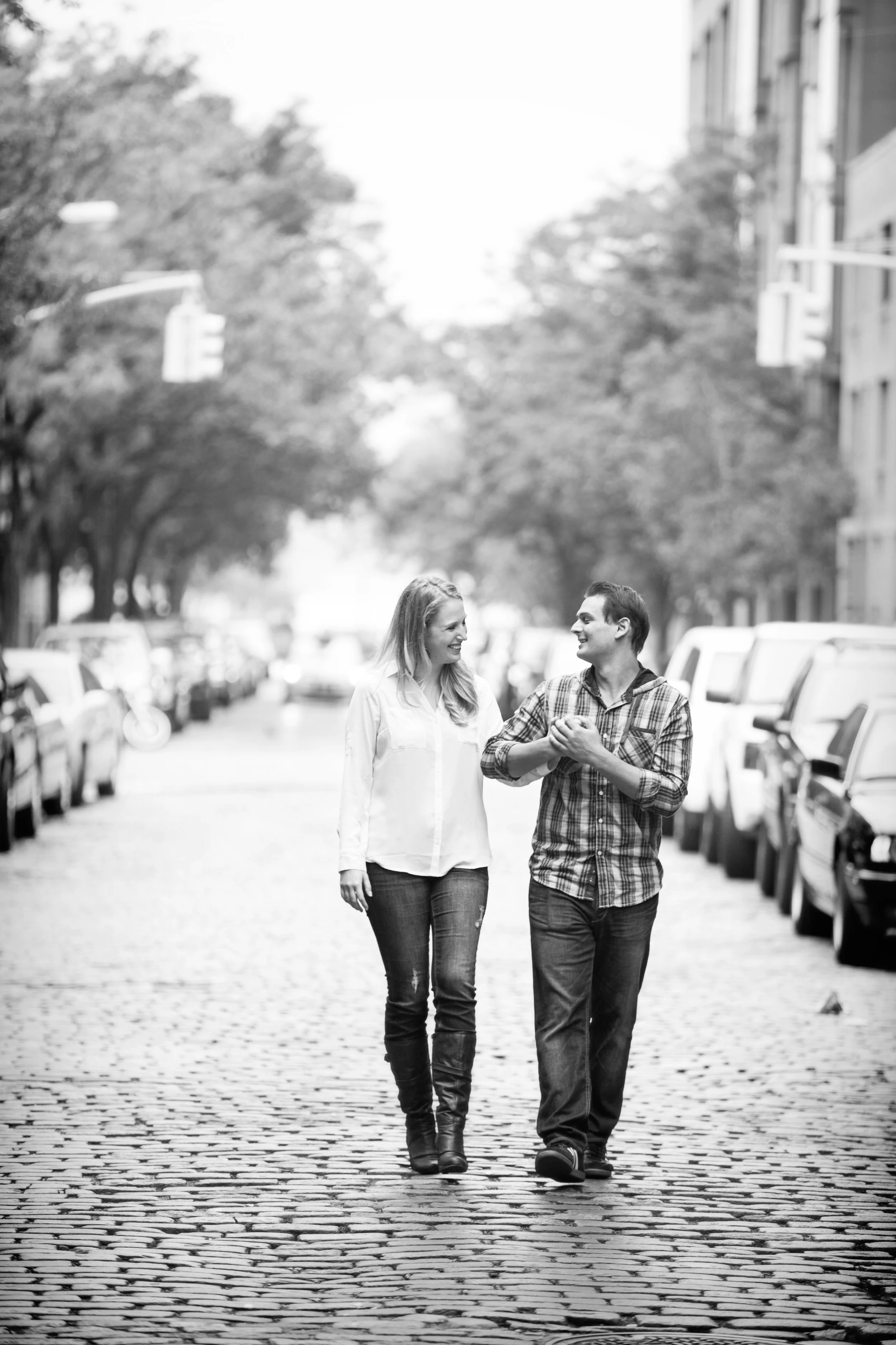 A black and white photo of a couple walking hand in hand on a cobblestone street, smiling and looking at each other, with parked cars and trees in the background.