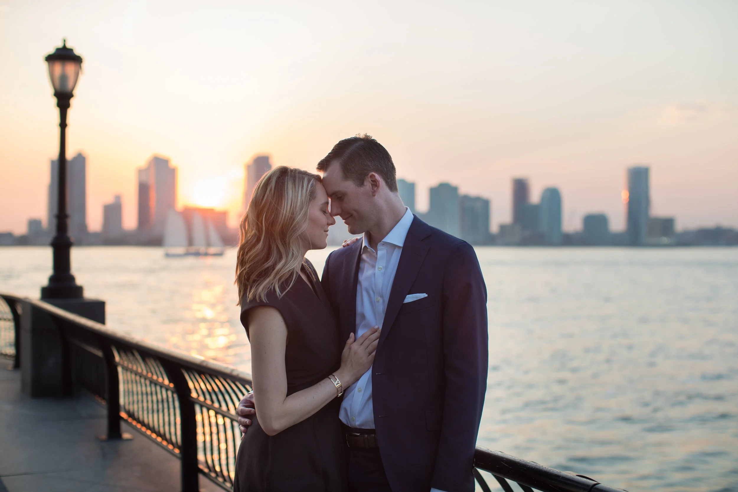 A couple standing close together by the water's edge during sunset in a city, with their foreheads touching and eyes closed.
