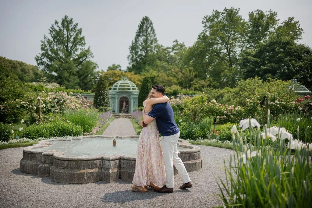 A couple hugging in a garden with a fountain and lush green trees in the background.