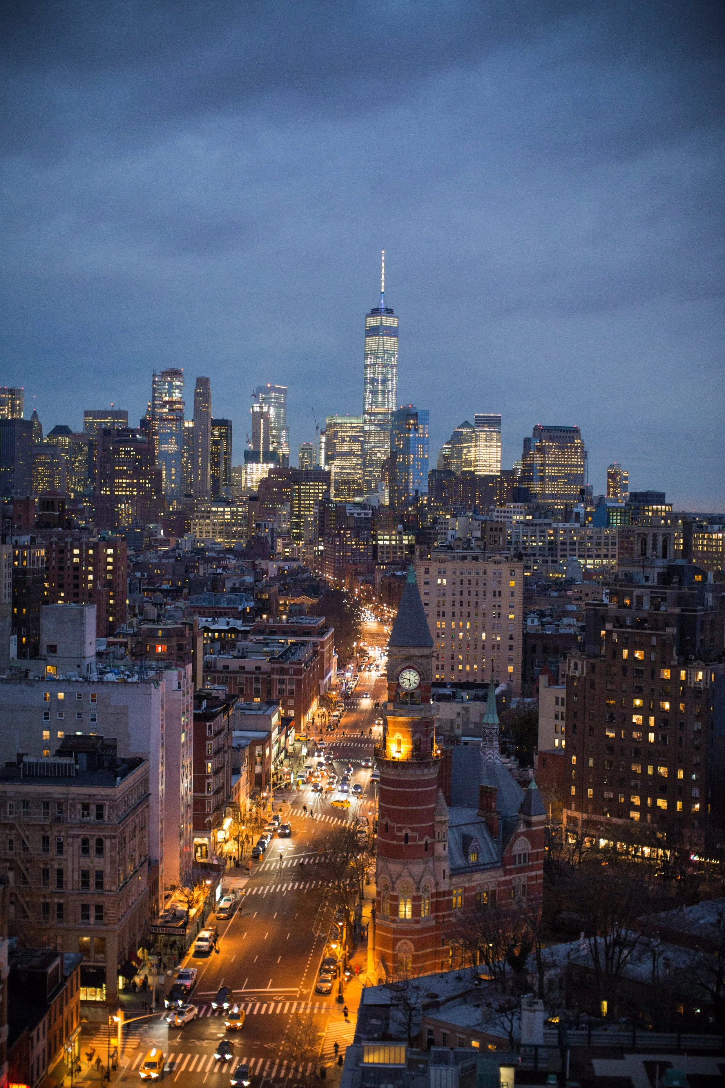 Nighttime cityscape of Manhattan, New York City, with illuminated buildings including One World Trade Center, and a historic clock tower on a corner, with city streets and traffic illuminated by streetlights.