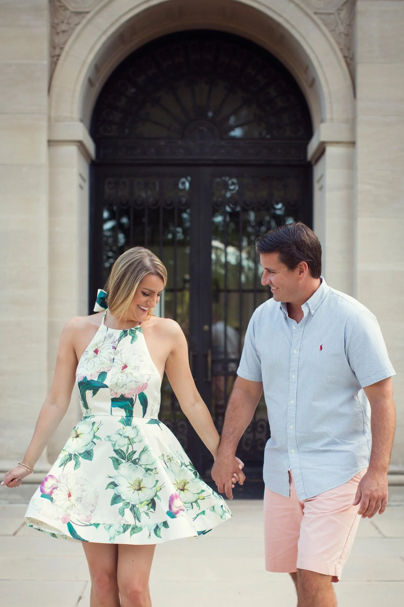 A young woman in a floral dress and a young man in a light blue shirt and pink shorts holding hands and smiling outside in front of a large black metal gate.