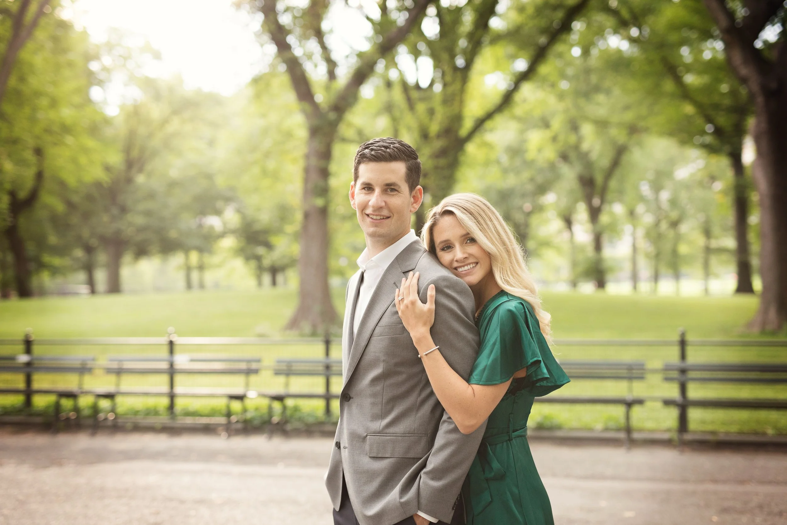 A smiling man in a gray suit and a woman in a green dress standing close together outdoors in a park with trees and benches in the background.