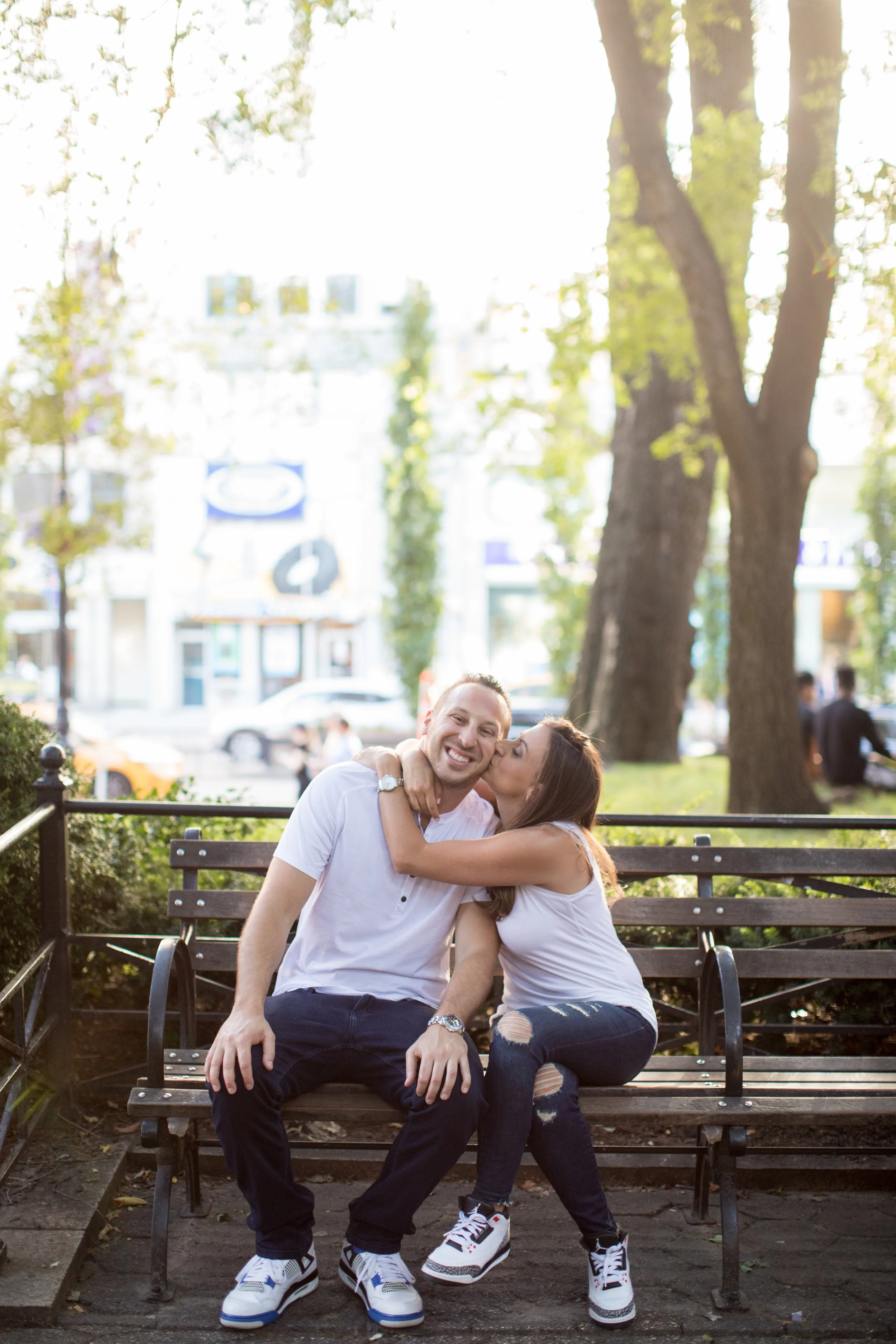 A man and a woman sitting on a park bench, with the woman kissing the man on the cheek. The man is smiling, wearing a white t-shirt, dark jeans, and sneakers. The woman is also wearing a white top, ripped jeans, and sneakers. They are surrounded by t