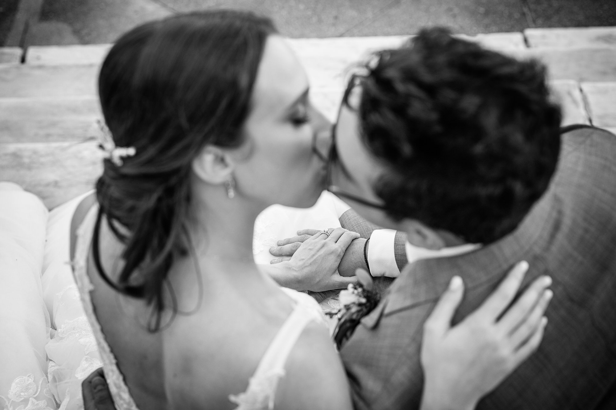A black and white photo of a couple kissing, with the woman wearing a lace dress and the man wearing a suit, on a wooden bench.