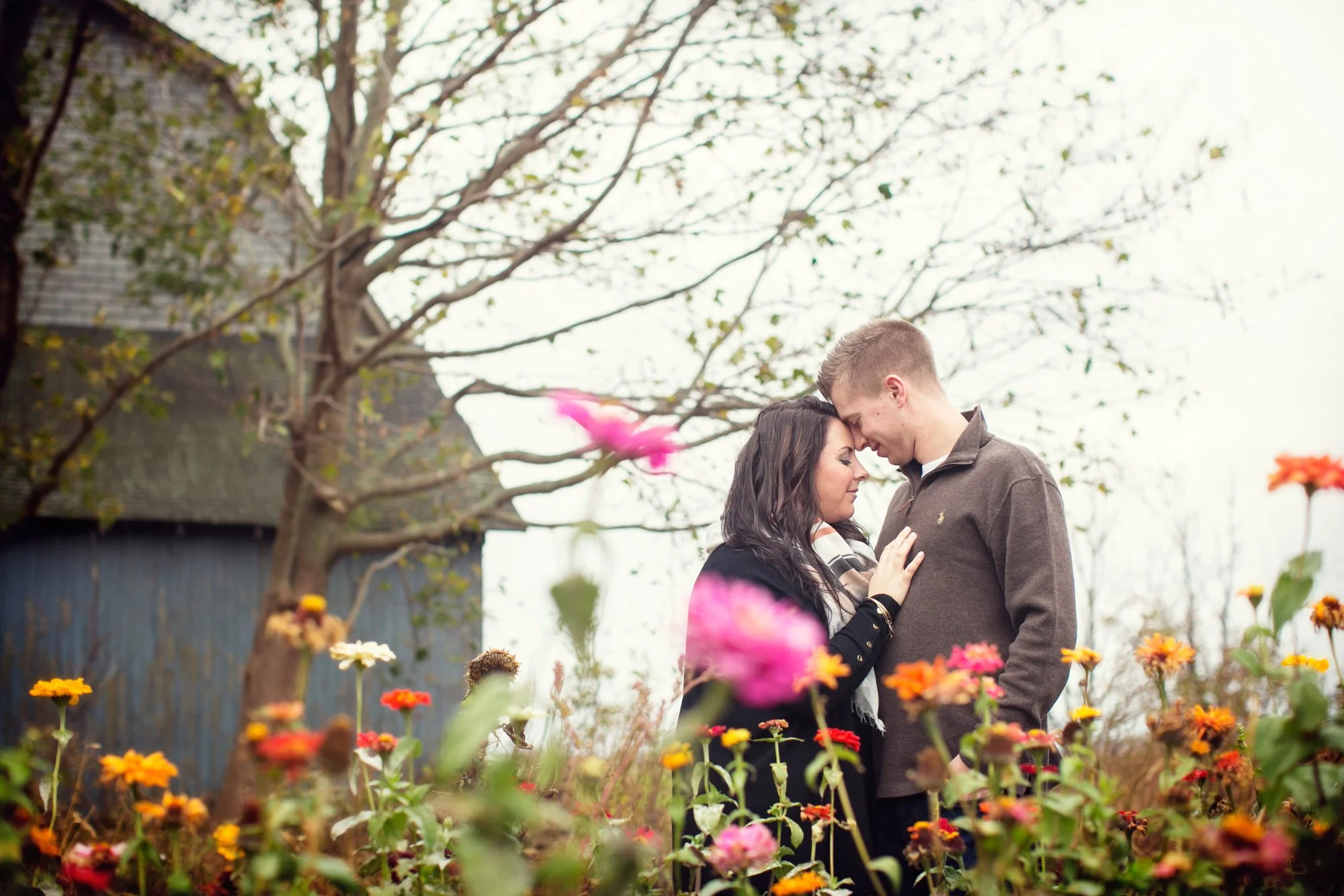 A couple standing close with foreheads touching in a garden with colorful flowers and a tree in the background.