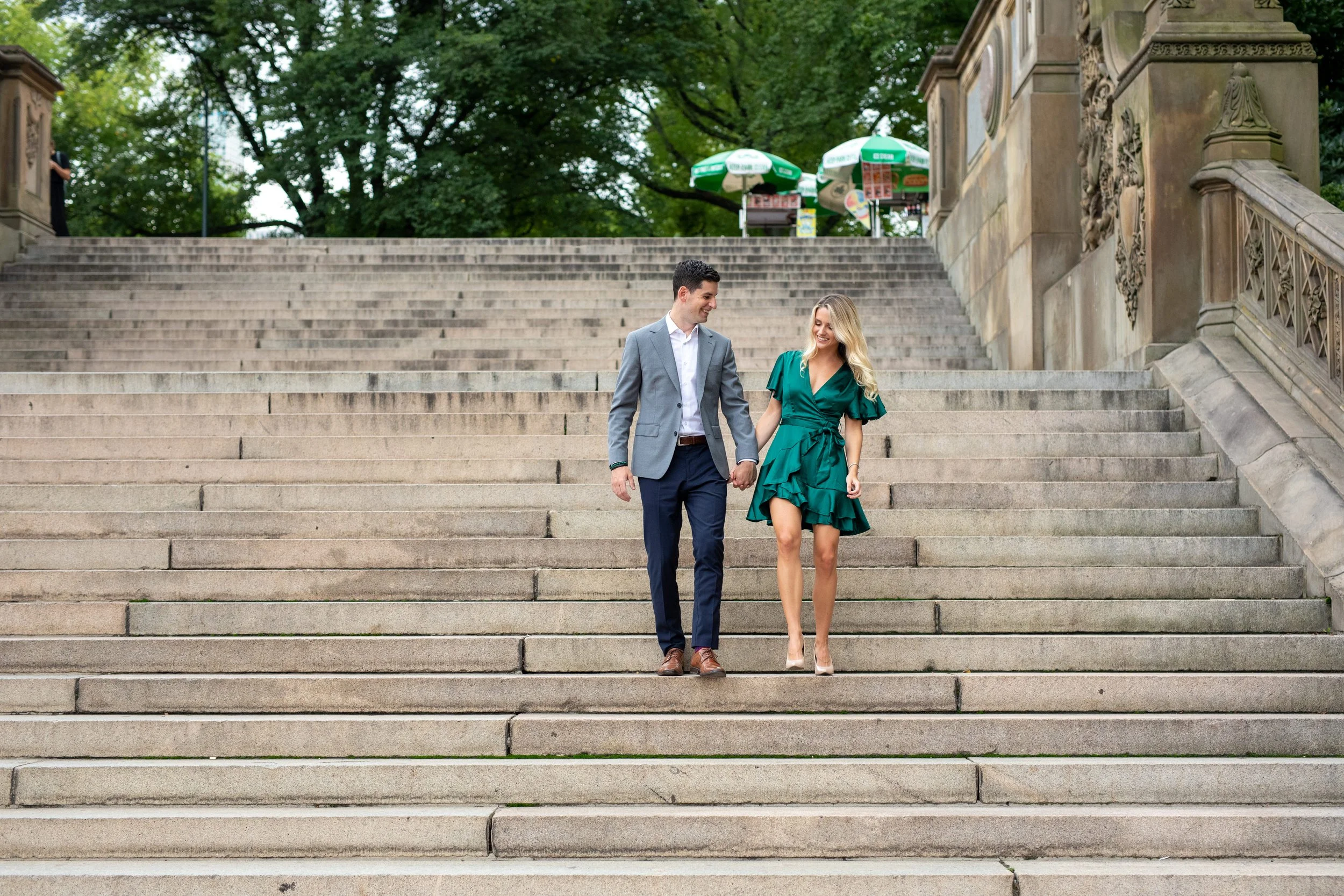 A couple walking down a large outdoor concrete staircase, holding hands and smiling at each other, with trees and umbrellas in the background.