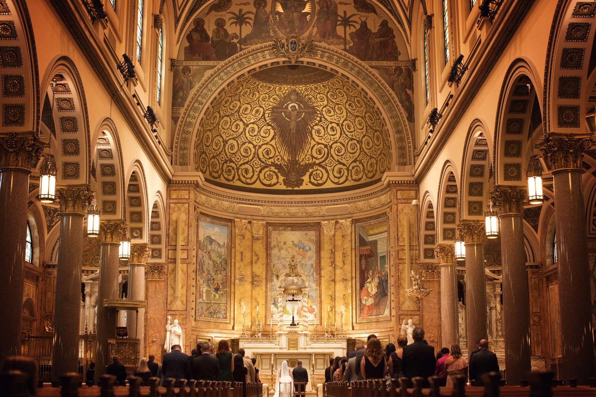 People praying inside a grand cathedral with ornate gold and marble decor, large paintings, and a high, domed ceiling with religious artwork, including a central figure with arms outstretched.