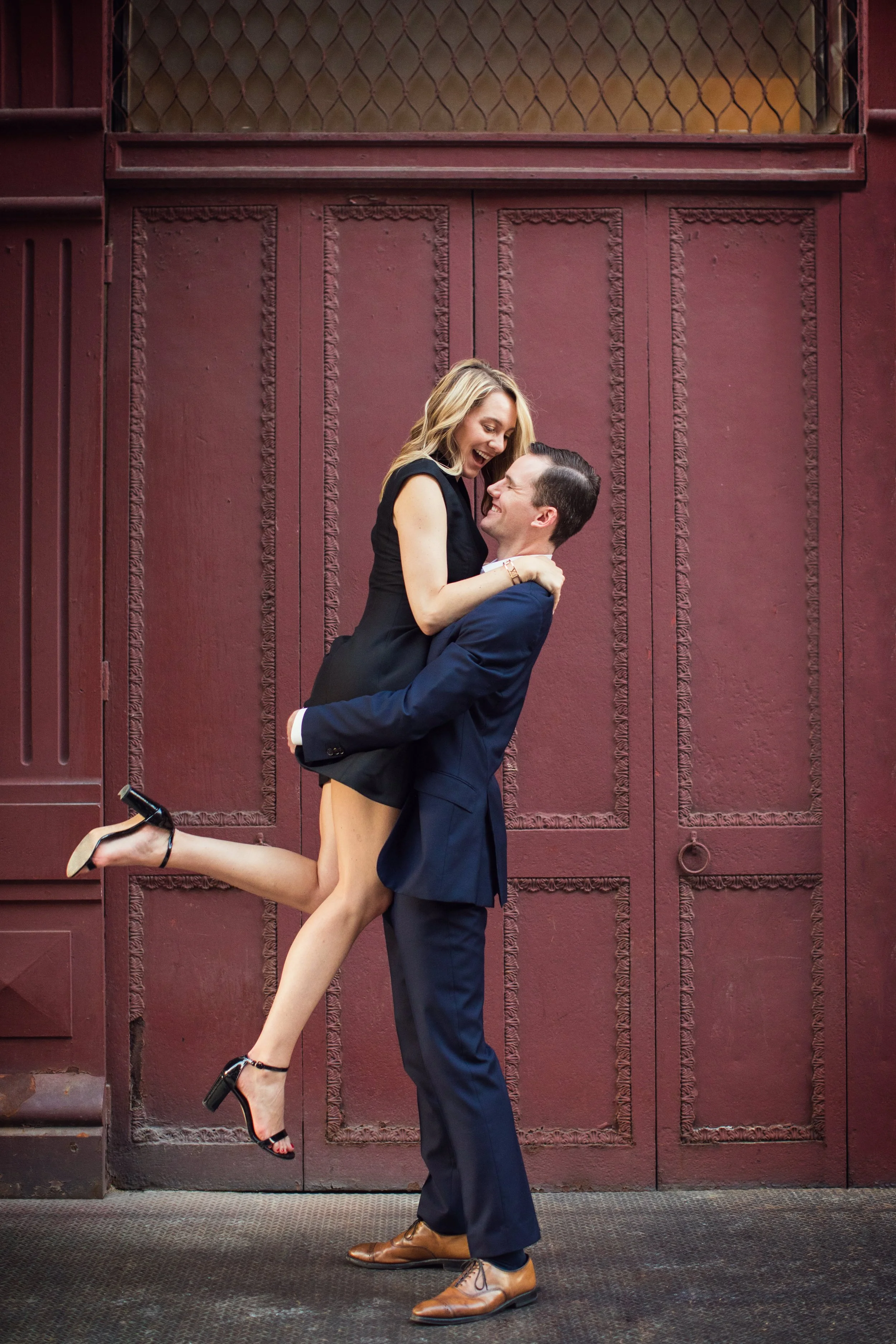 A man in a blue suit lifting a woman in a black dress and heels, both smiling and laughing in front of a burgundy wall.