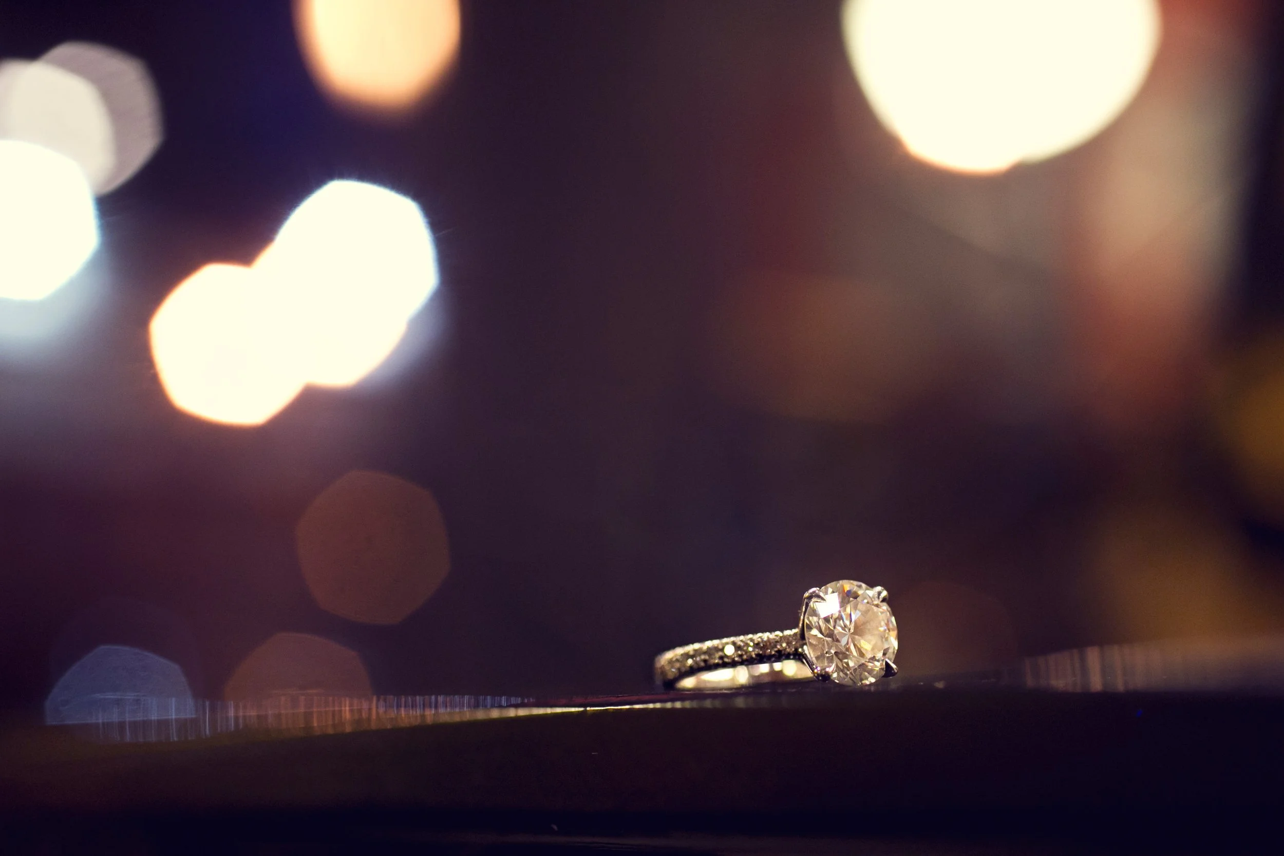 Close-up of a diamond engagement ring with a large central stone and a band encrusted with smaller diamonds, placed on a dark surface with blurred city lights in the background.