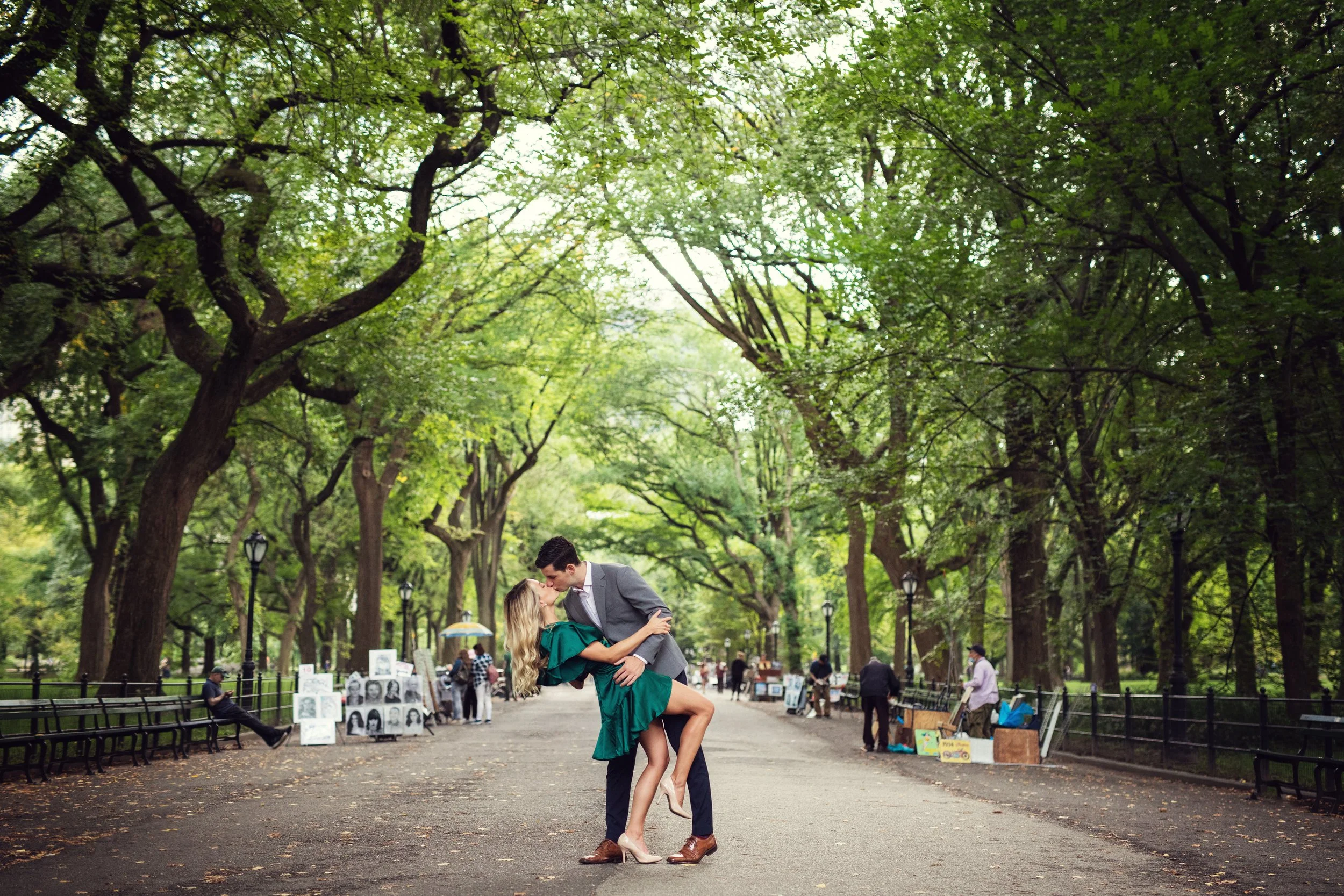 Couple kissing in a park with vendors selling artwork and street performers in the background.