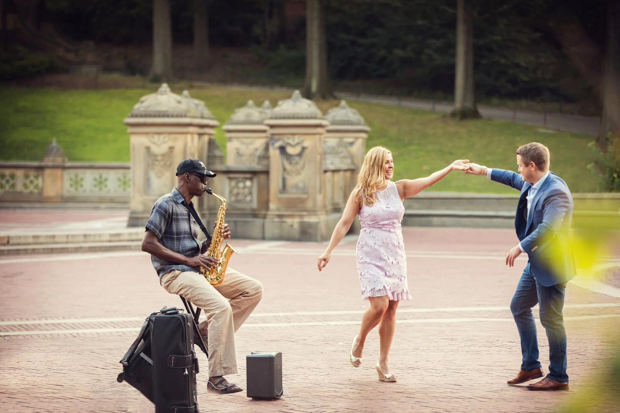 Couple dancing outdoors with a saxophonist playing nearby, on a brick paved area surrounded by greenery and historic stone structures.