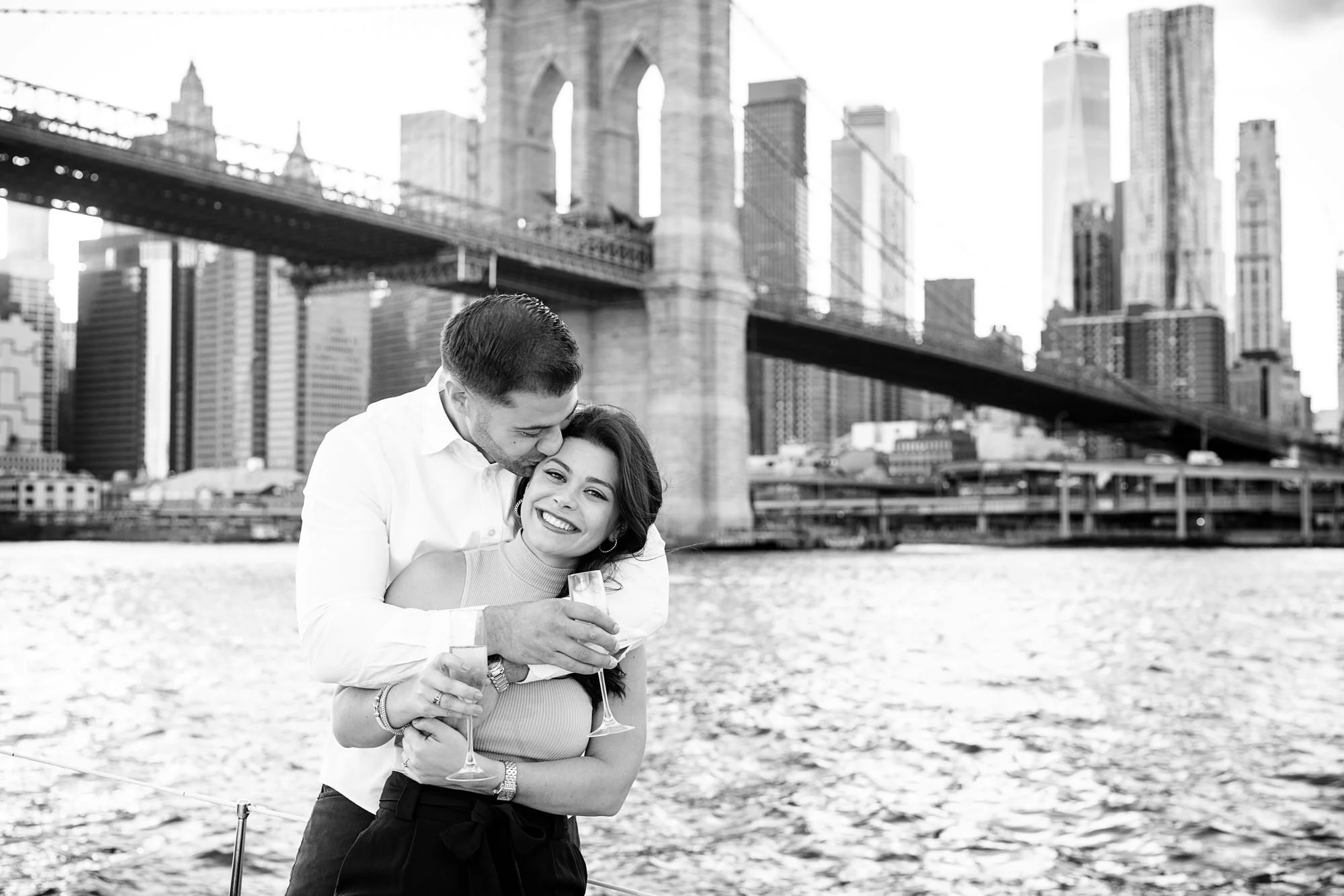 A couple enjoying drinks by the water with the Brooklyn Bridge and Manhattan skyline in the background.