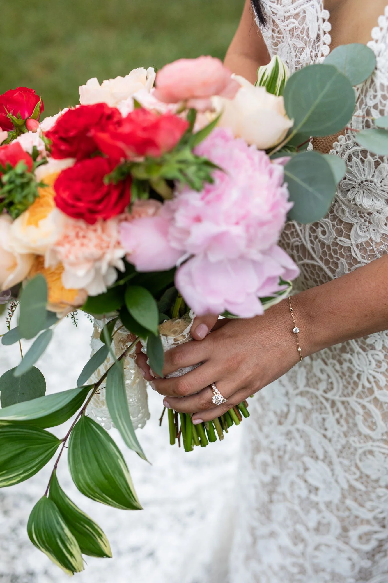 Close-up of a bride holding a bouquet of pink, white, red, and peach flowers with green leaves, wearing a lace dress, ring, and bracelet.