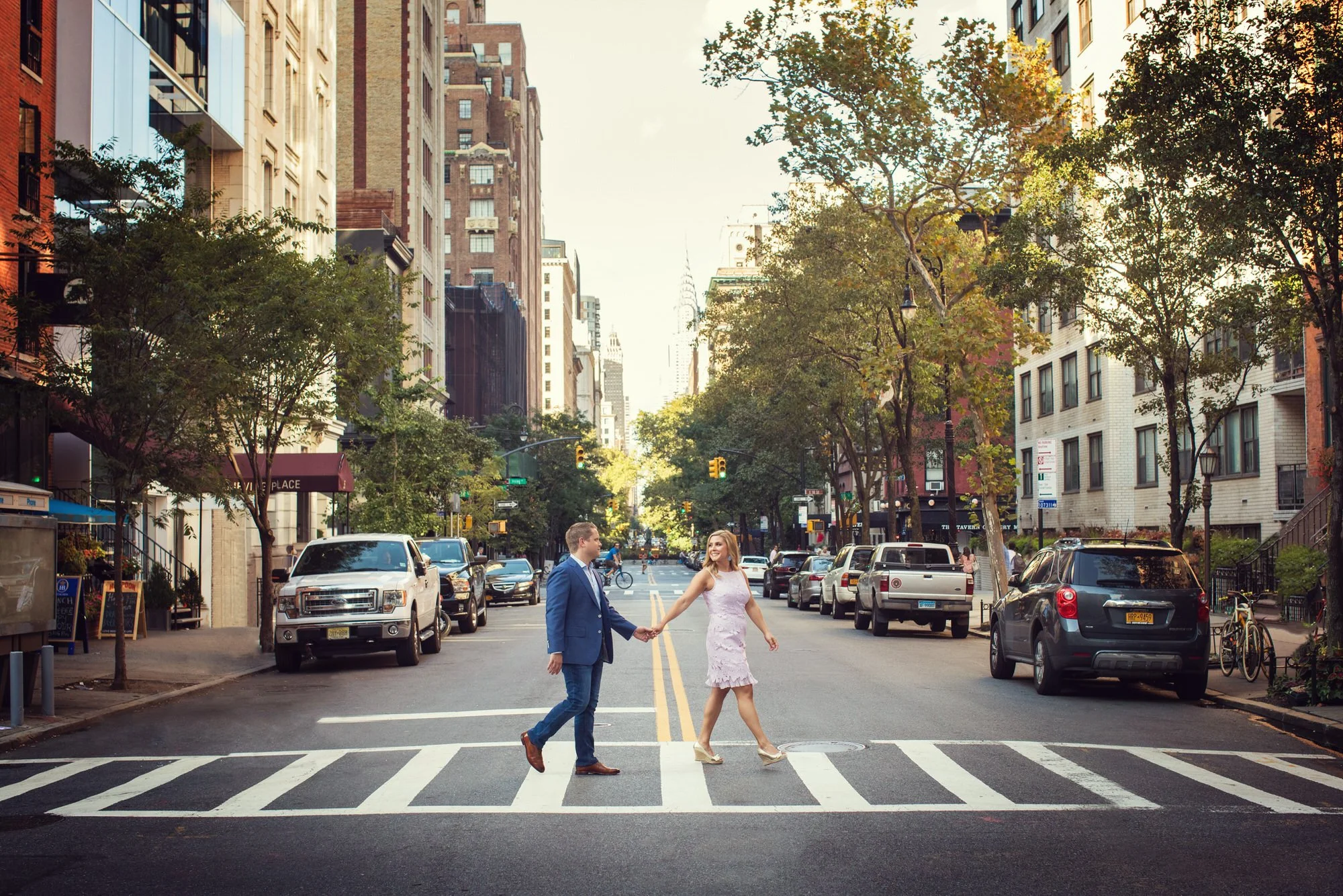 A man and woman holding hands and walking across a crosswalk in an urban city street, surrounded by parked cars, trees, and tall buildings, with the man dressed in a blue suit and the woman in a light pink dress.