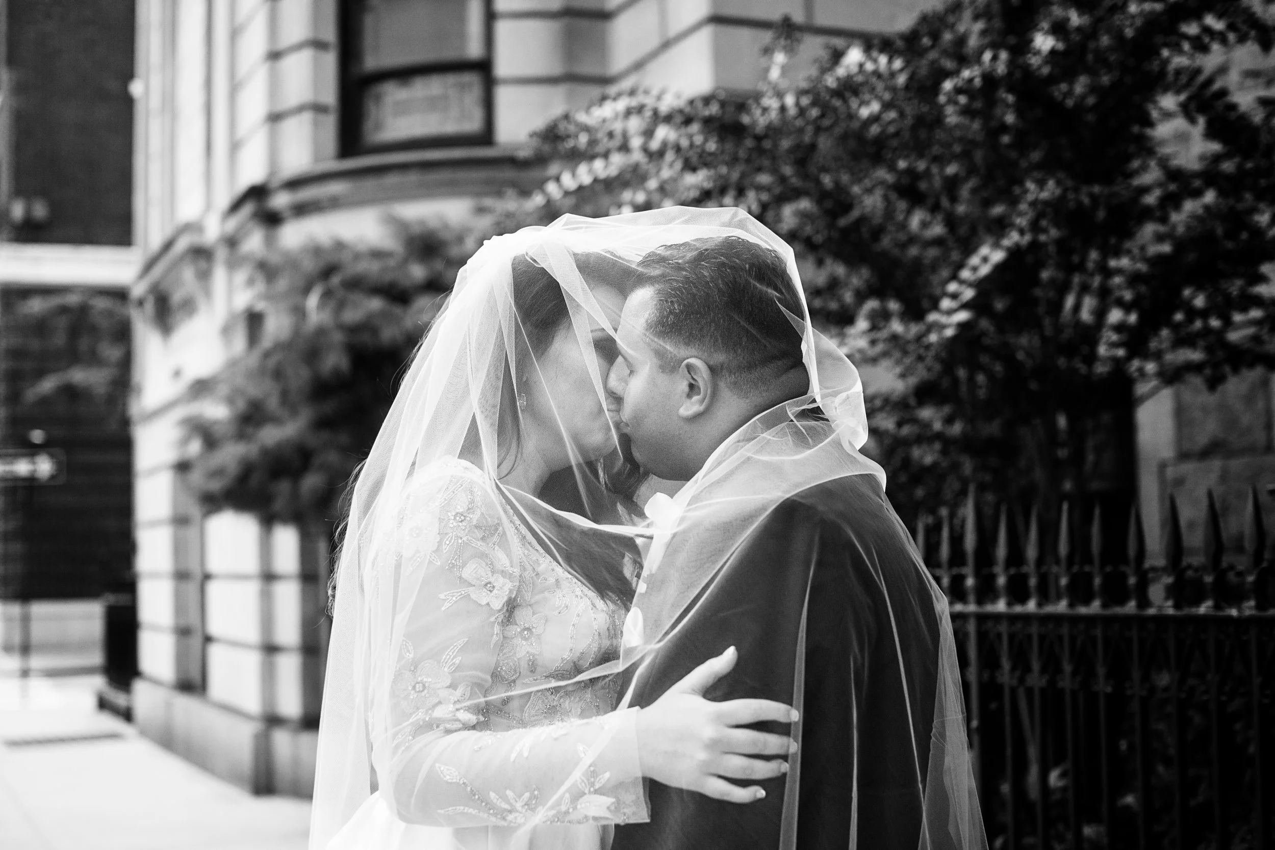 A black and white photo of a bride and groom sharing a kiss under the bride's veil outdoors in front of a building with trees and a fence.