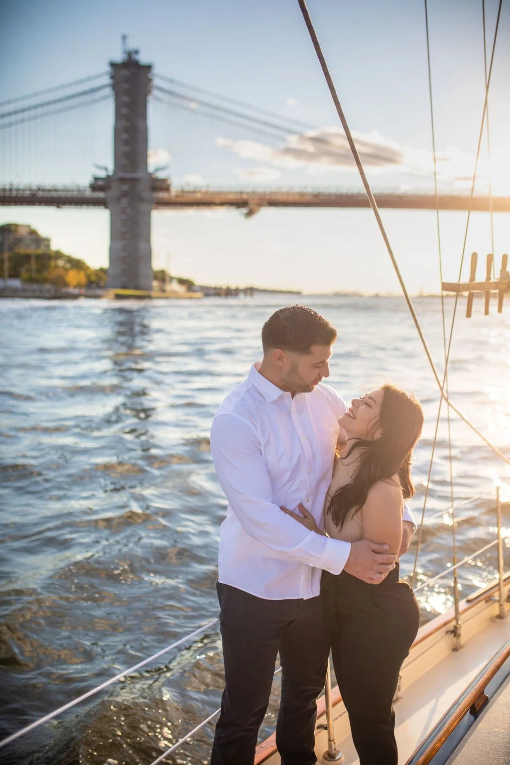 A couple stands close together on a boat, smiling and looking at each other, with a bridge and water in the background during sunset.