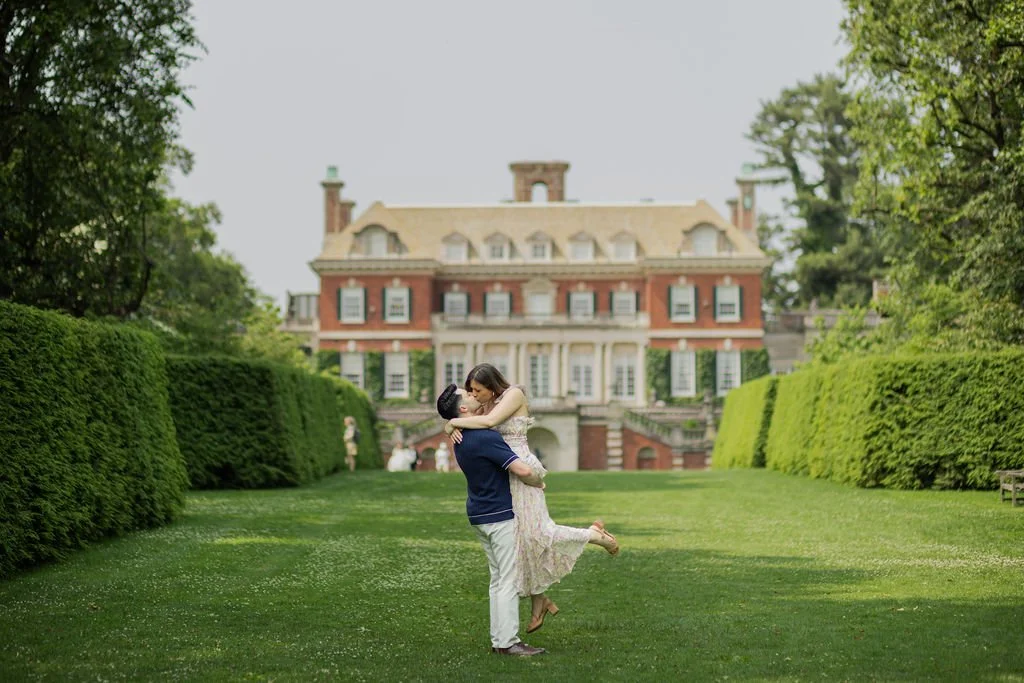 A couple is dancing on a manicured lawn in front of a large historic brick mansion with a yellow roof, surrounded by green trees and bushes.
