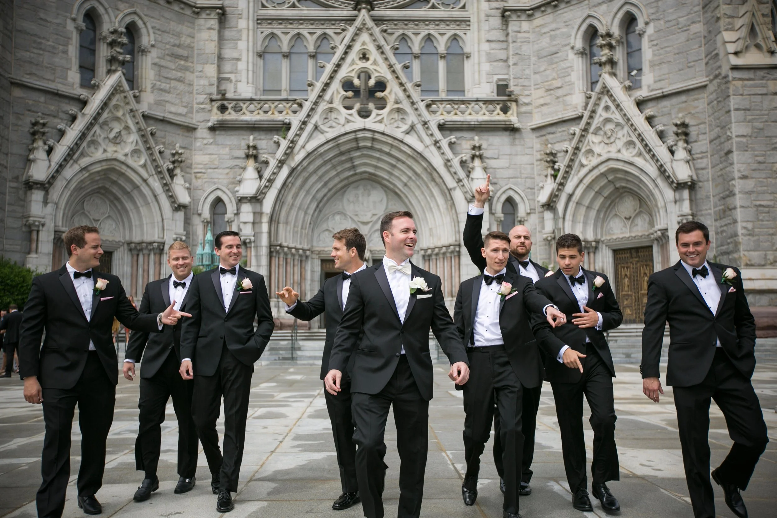 A group of men in tuxedos with boutonnières, smiling and posing playfully outside a large Gothic-style cathedral with ornate stone architecture.