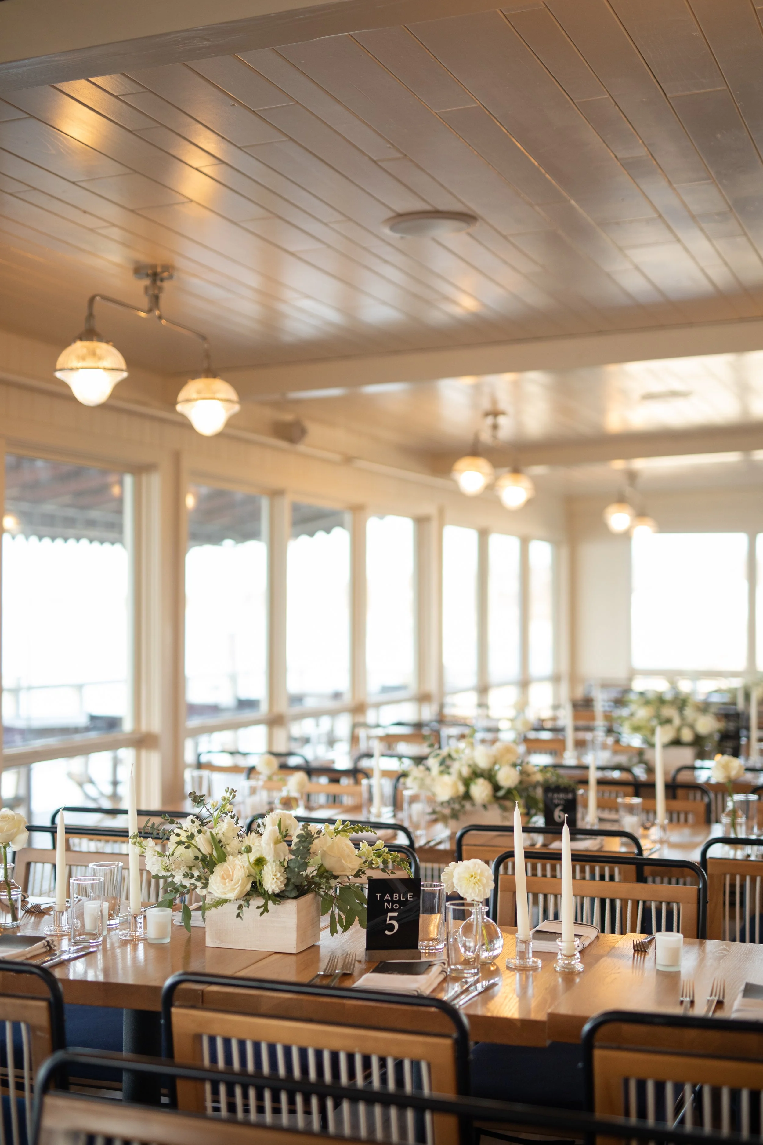 A decorated banquet hall with wooden tables, white floral centerpieces, and tall white candles. The tables are set with glassware and silverware, with a table number sign for table 5. Large windows line the side of the room, allowing natural light to