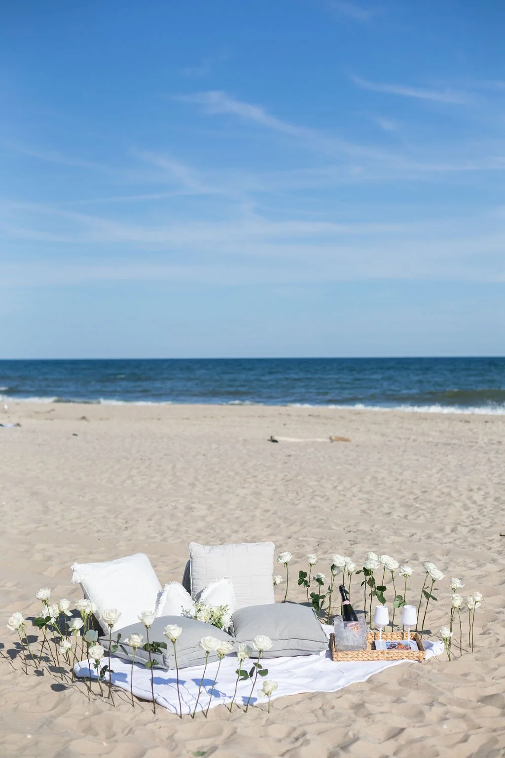 A beach scene with a white blanket and pillows on the sand, decorated with white roses, and a tray with glasses, a wine bottle, and snacks, overlooking the ocean under a blue sky.