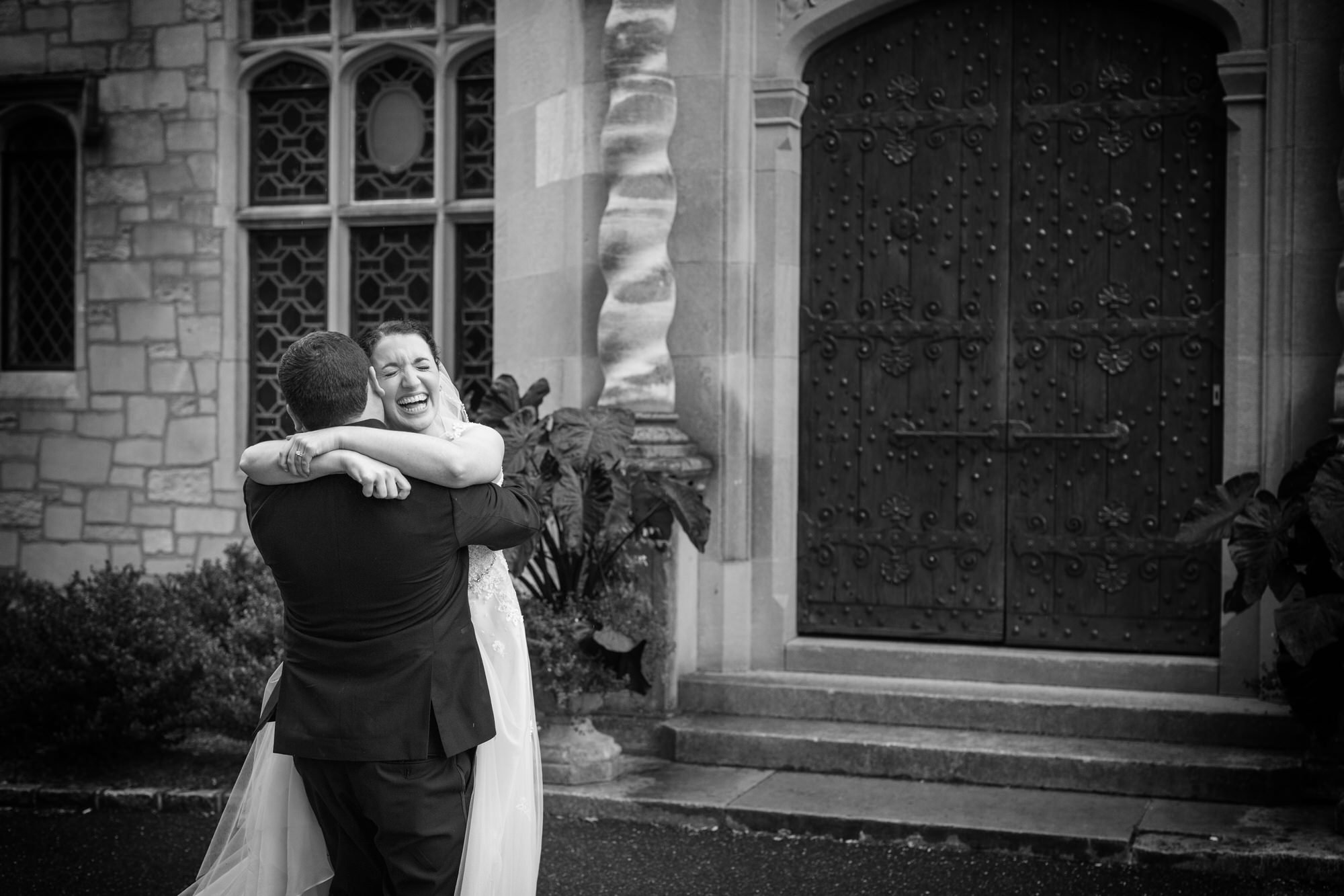 A bride and groom embrace outside a historic building with large ornate double doors, stone steps, and garden plants, celebrating their wedding.