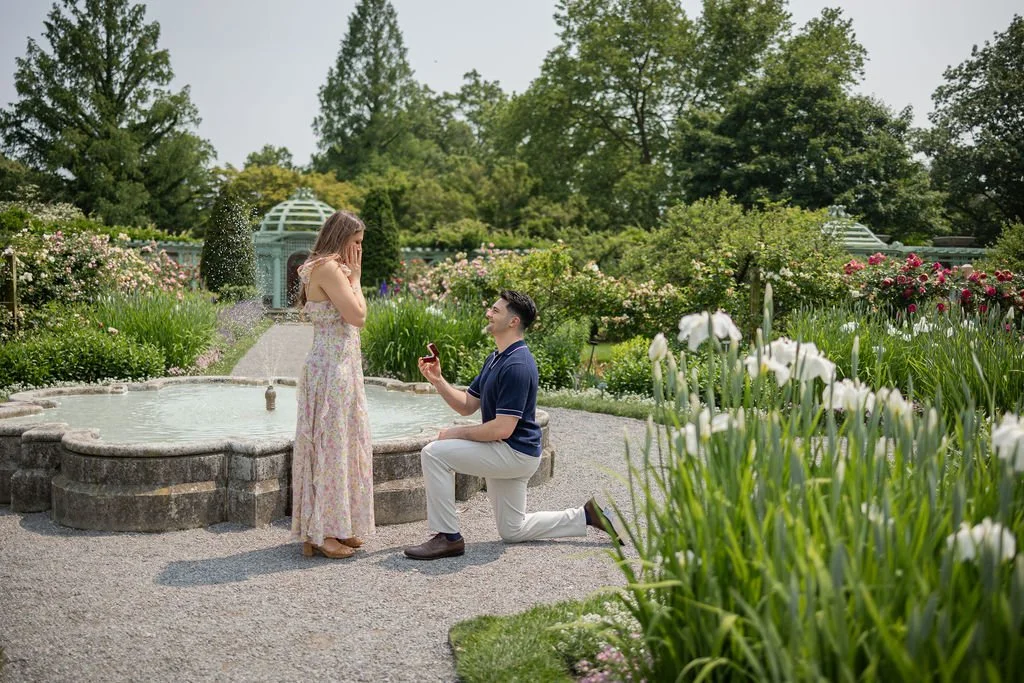 A man kneeling on one knee proposing to a woman in a garden with flowers, a fountain, and a glass greenhouse in the background.
