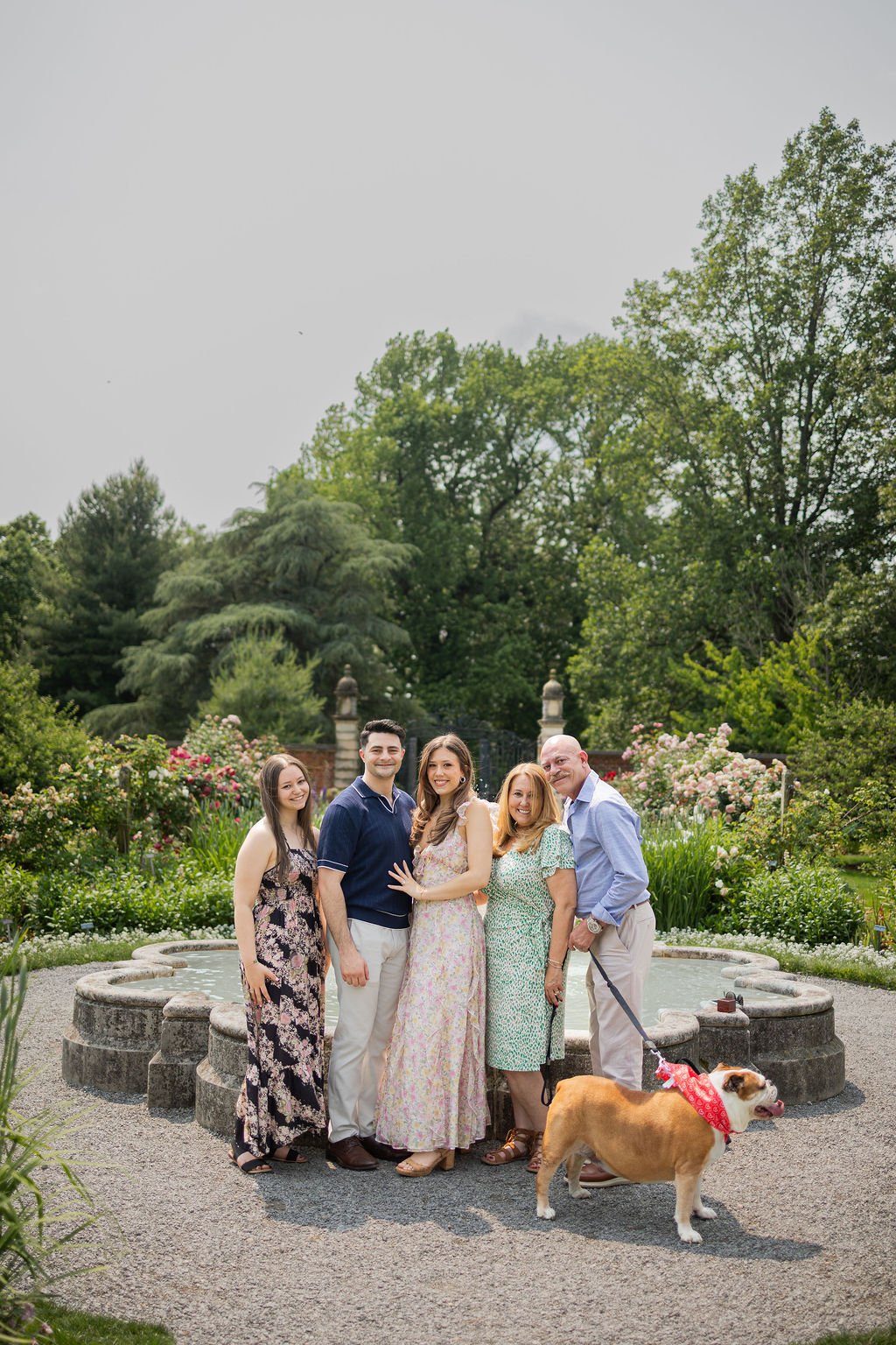 A group of five people and a dog standing in a garden with lush green trees and flowering bushes in the background.