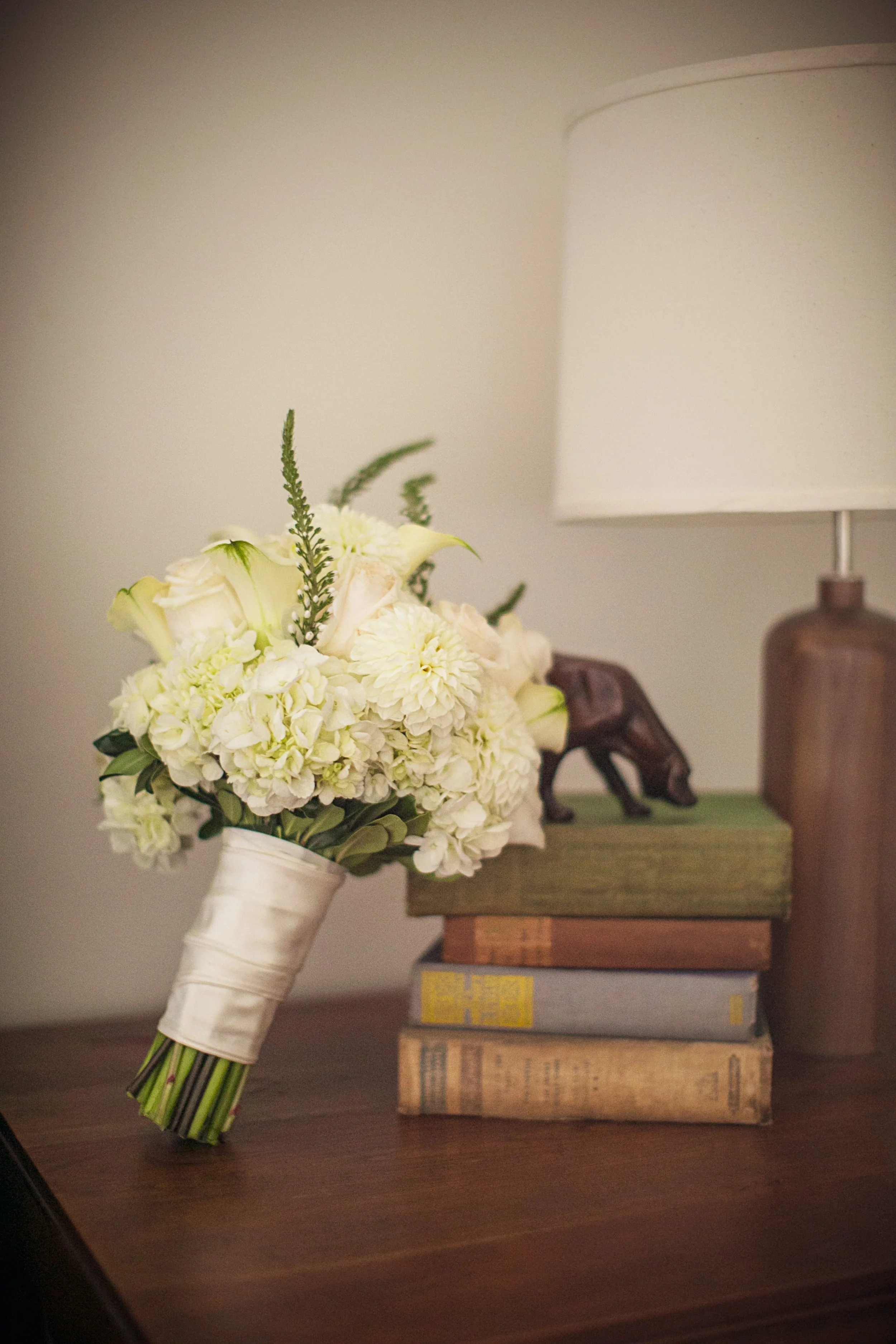 A bouquet of white flowers, including roses and dahlias, wrapped with a satin ribbon, placed on a wooden surface next to a stack of old books and a lamp with a beige shade.