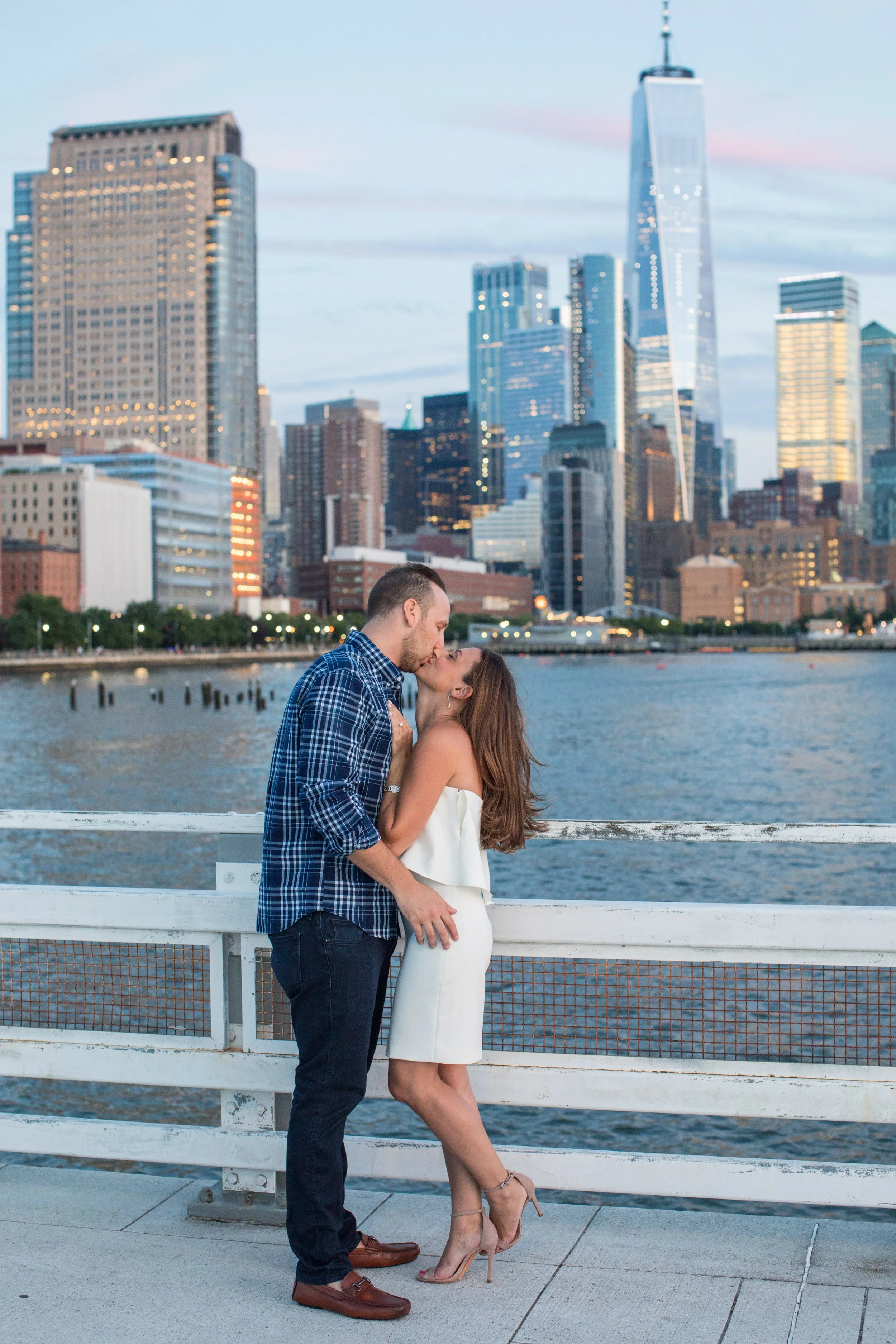 A couple kissing on a waterfront promenade with the New York City skyline, including One World Trade Center, in the background during sunset.