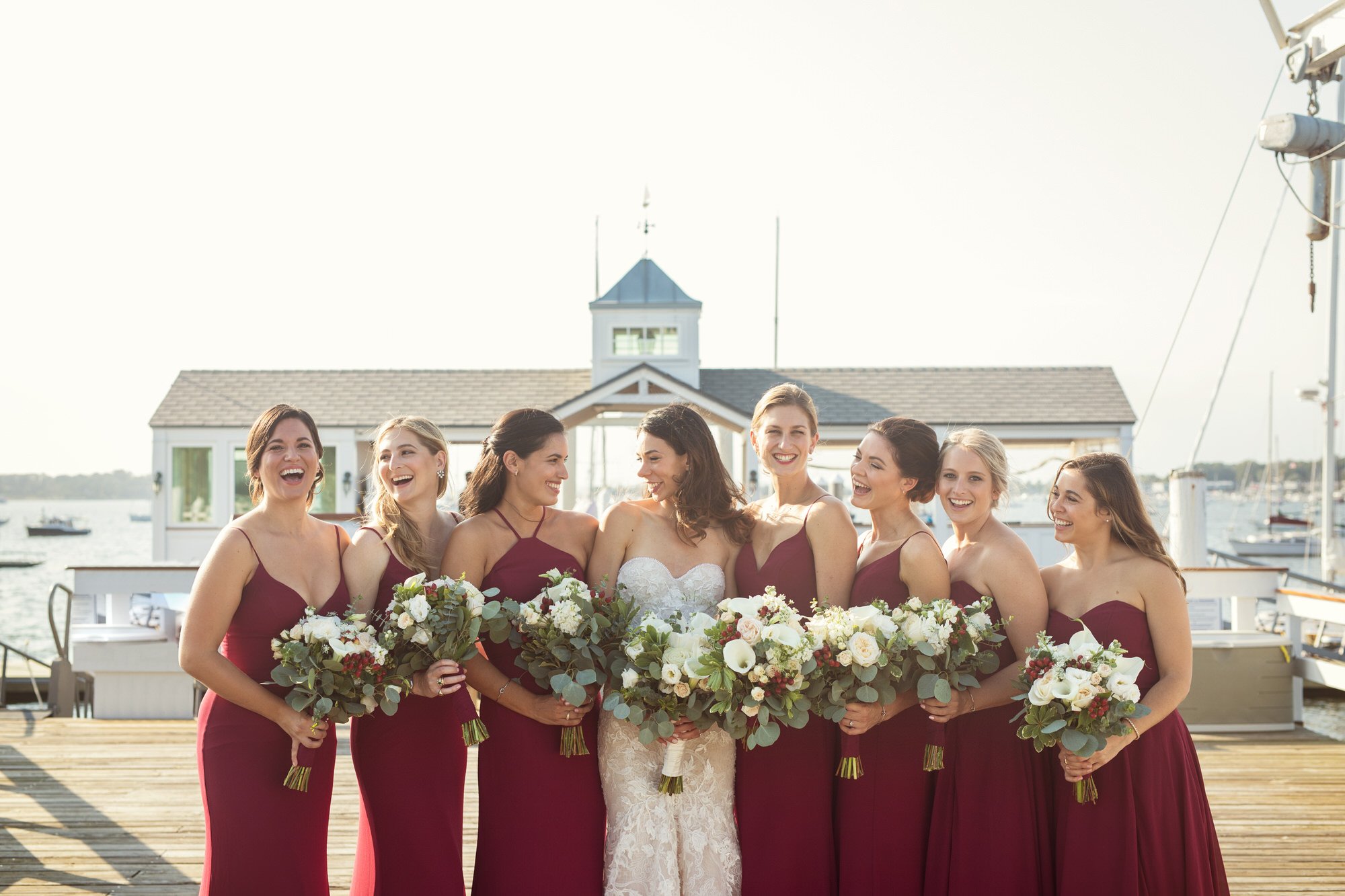 Bride and bridesmaids in burgundy dresses holding bouquets, standing on a dock near a marina with boats, during daylight.
