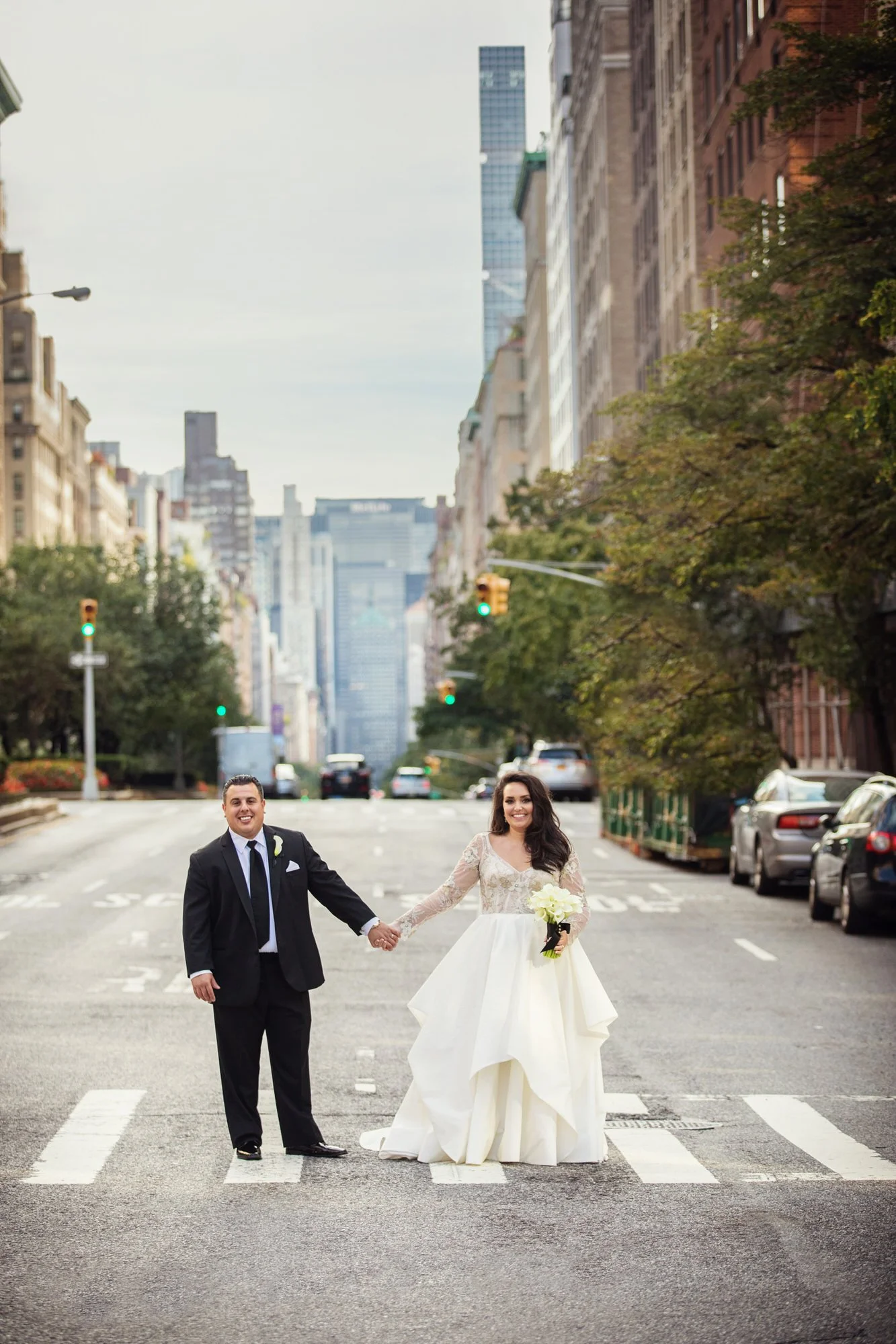 A newlywed couple holding hands and smiling on a city street at the crosswalk, the bride in a white wedding gown holding a bouquet of white roses, and the groom in a black suit, with tall buildings and parked cars in the background.