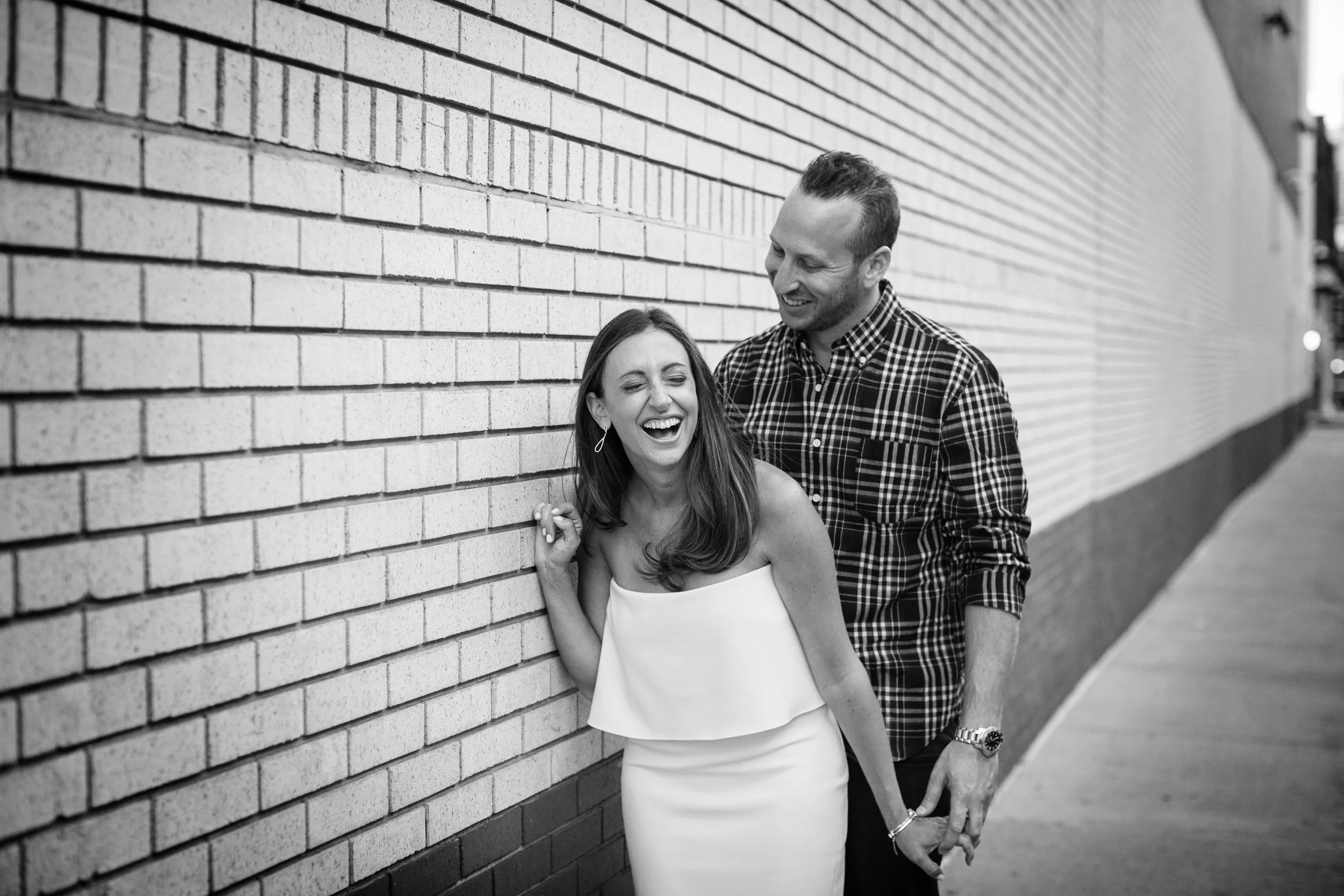 A happy couple, a woman in a white dress and a man in a checkered shirt, laughing and holding hands while leaning against a brick wall on a city sidewalk.