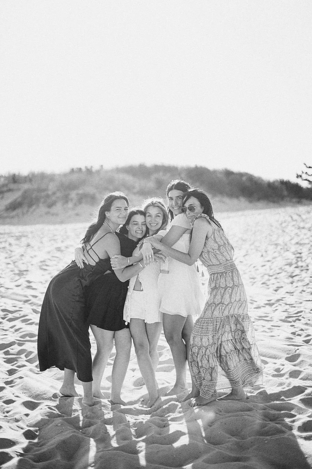 A group of six women hugging and smiling on a sandy beach during daytime, with a hill in the background, dressed in summer clothing.