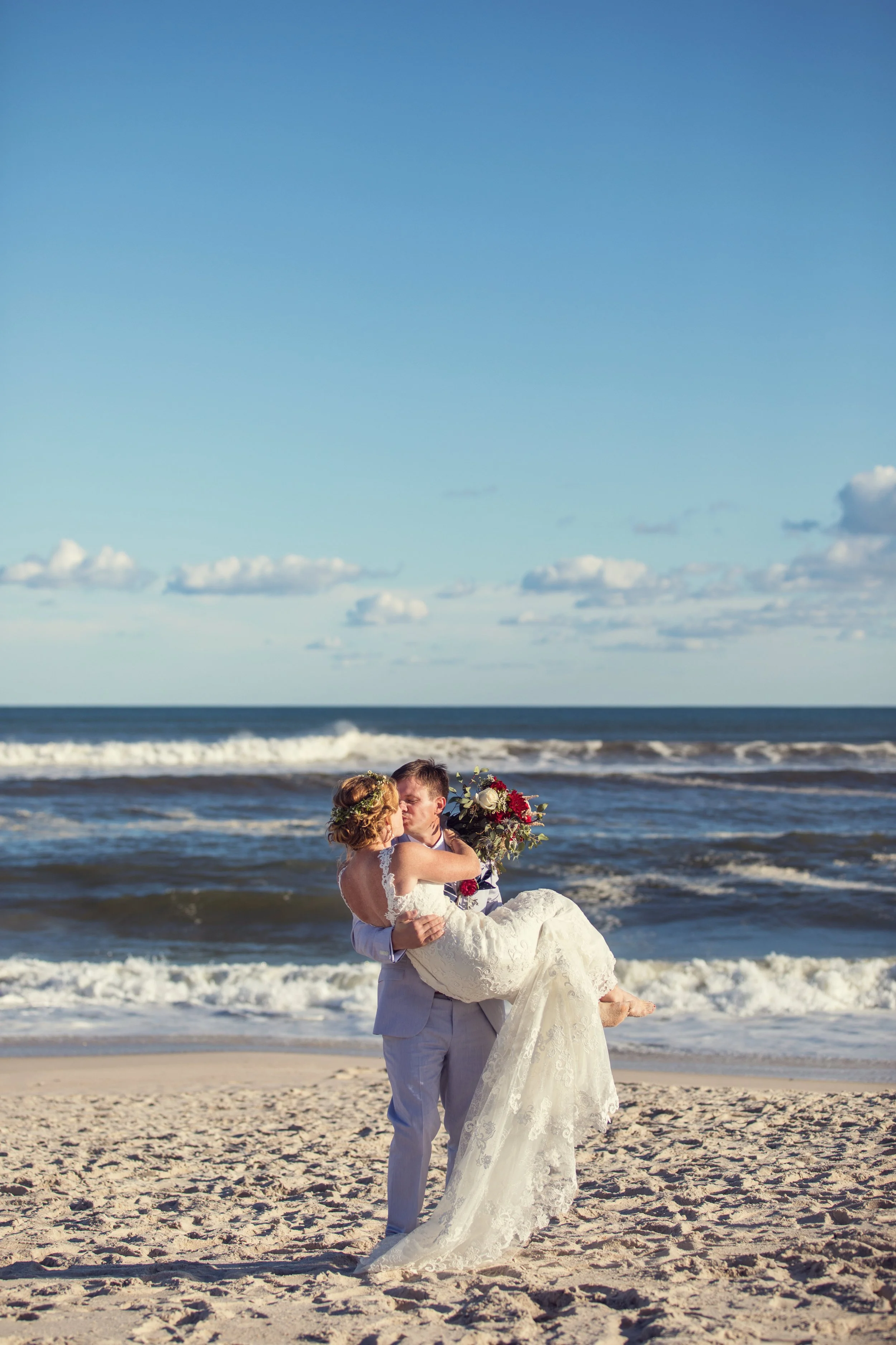 A newlywed couple sharing a kiss on the beach, with the groom holding the bride in his arms. The bride is wearing a lace wedding dress, and the groom is in a light-colored suit. The bride is holding a bouquet of flowers. The ocean waves and a blue sk
