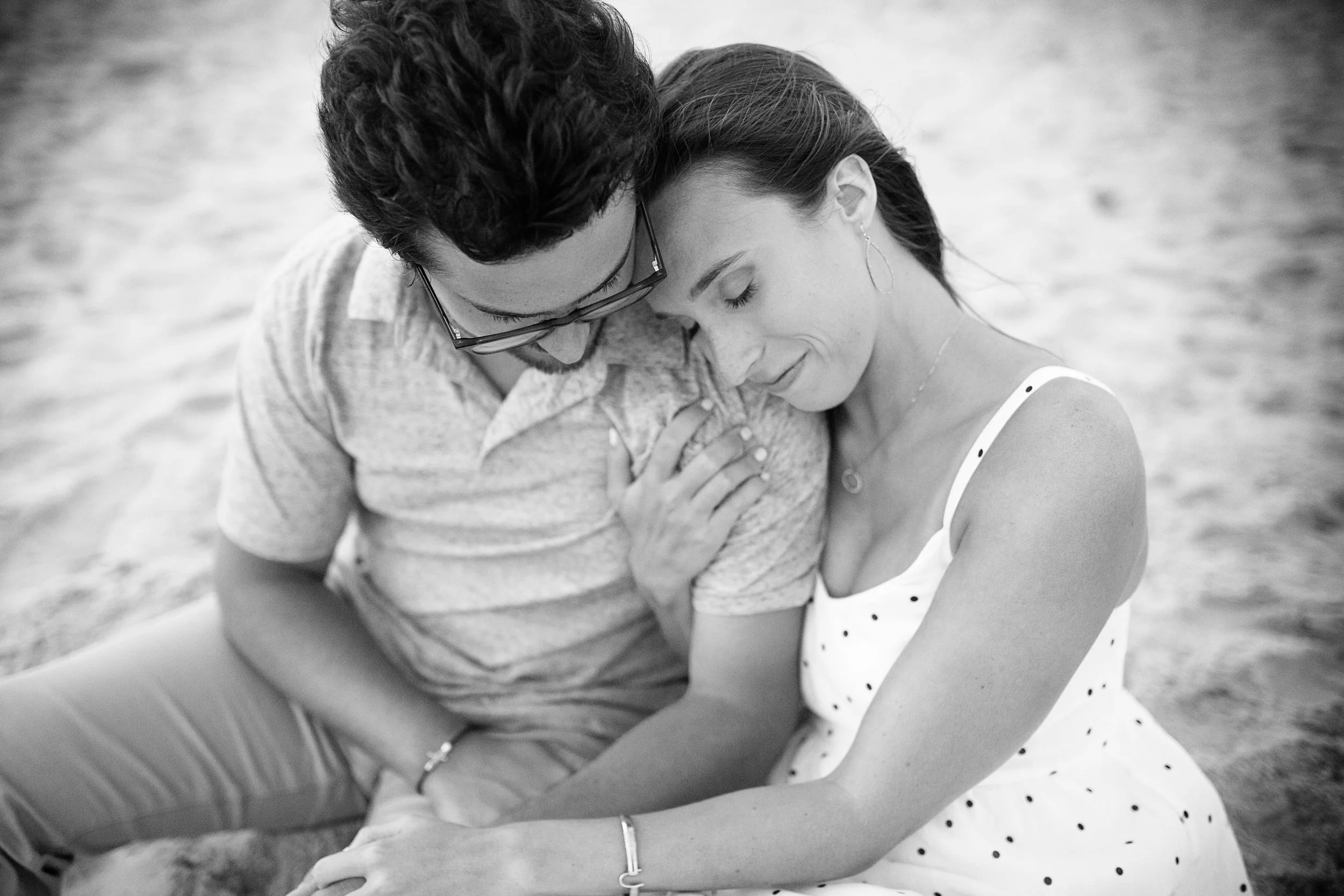A black-and-white photo of a couple sitting close together on the sand, with their foreheads touching and eyes closed, sharing an intimate moment.