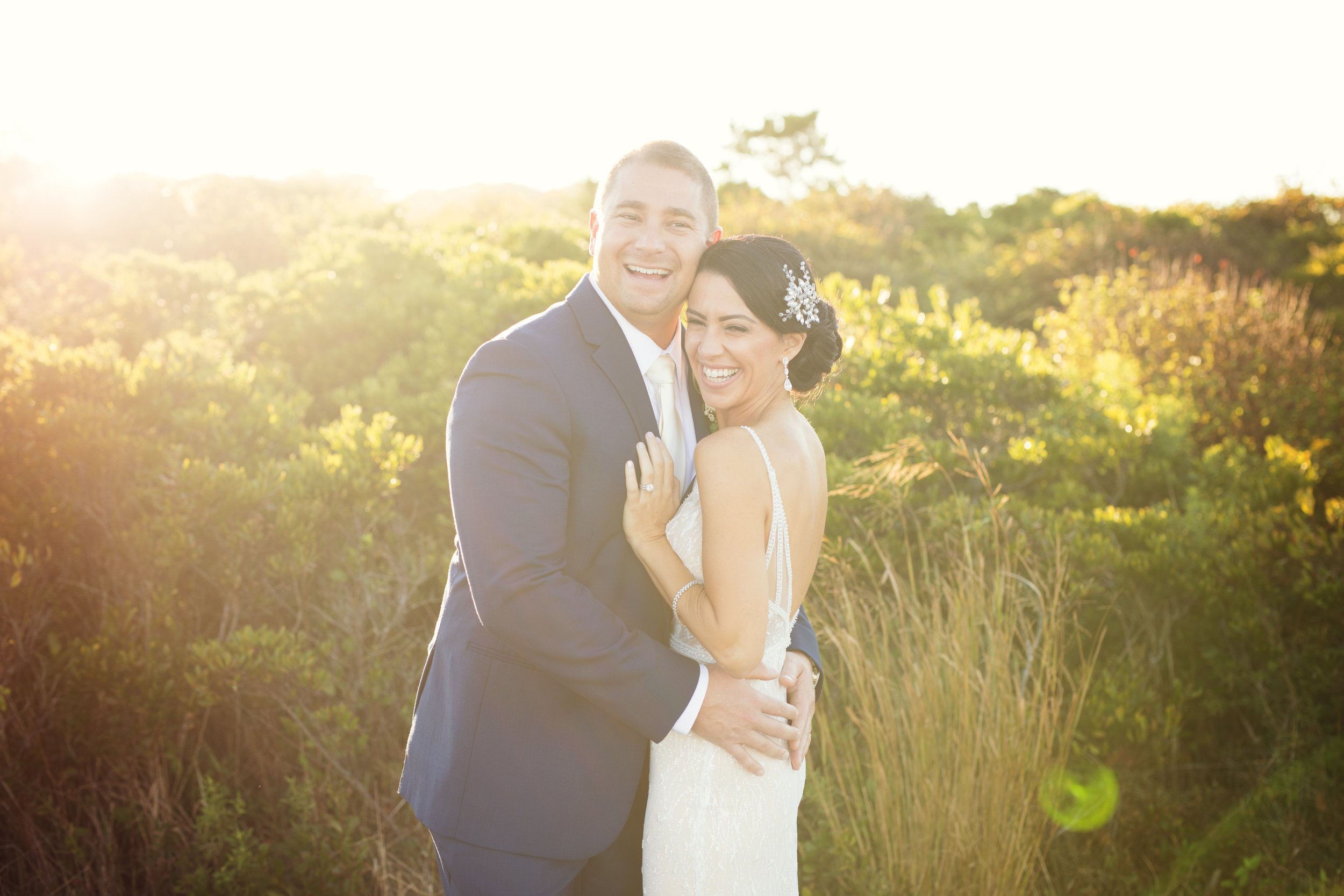 A newlywed couple in wedding attire happily embracing outdoors, with sunlight shining behind them and greenery in the background.