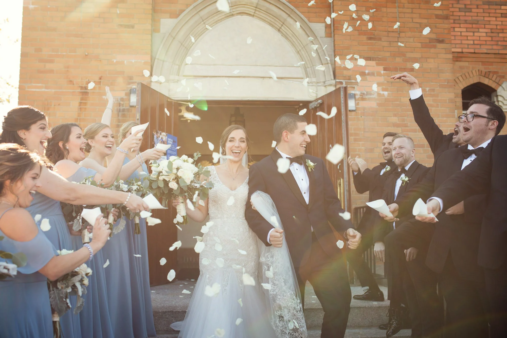 Bride and groom exiting a church under falling confetti, surrounded by smiling bridesmaids in blue dresses and groomsmen in tuxedos.
