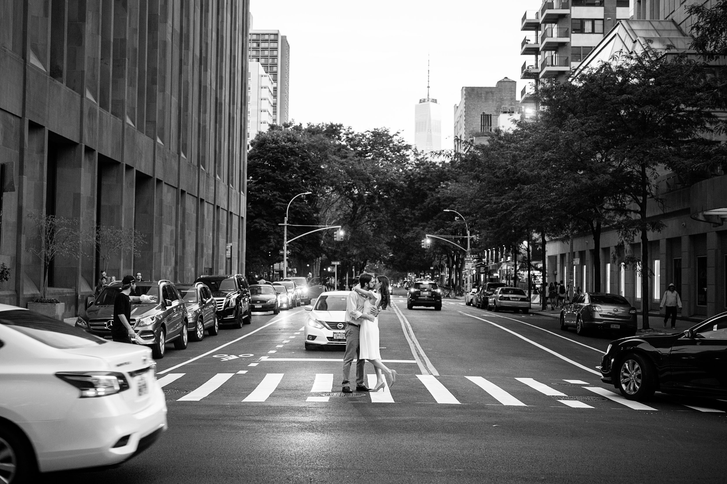 A black and white photo of a city street with a couple kissing in the crosswalk, surrounded by parked cars, buildings, and trees.