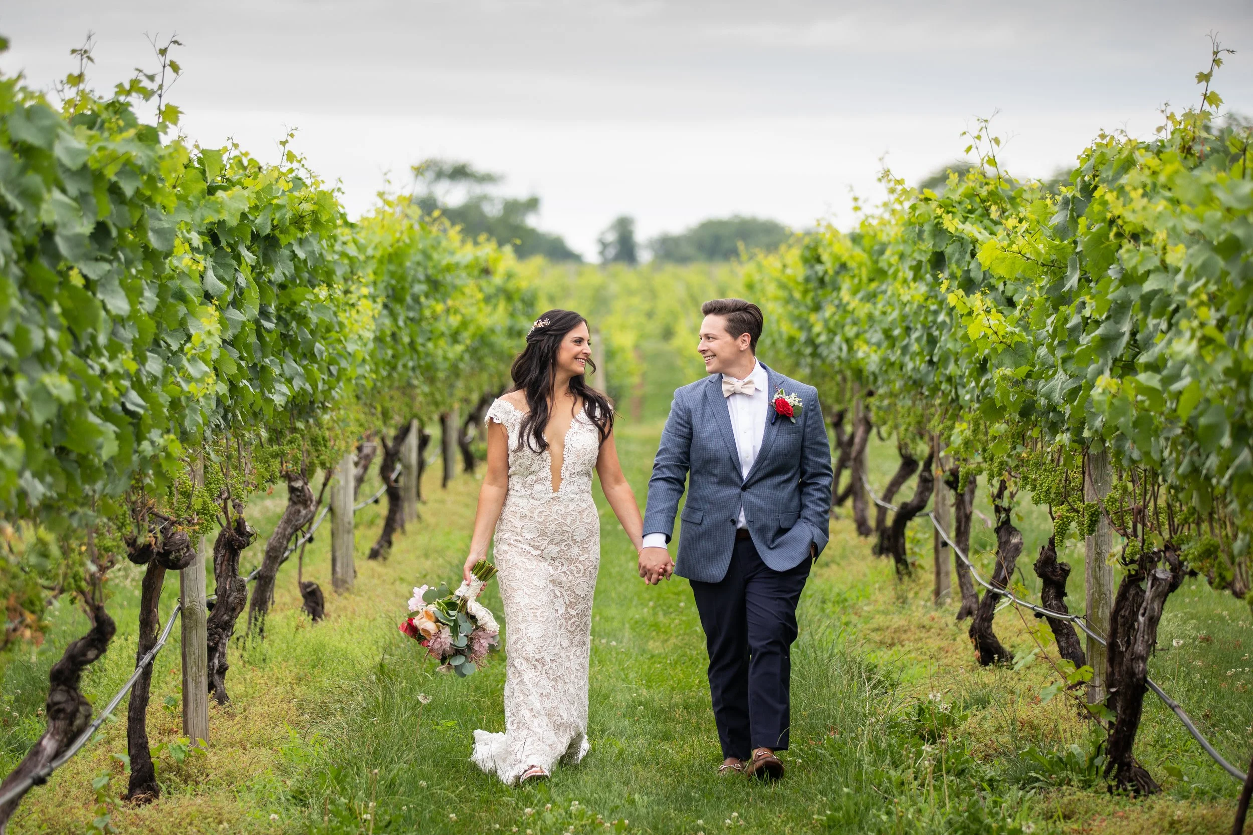 A bride and groom walking through a vineyard holding hands, smiling at each other. The bride is in a white lace wedding dress holding a bouquet, and the groom is in a blue suit with a white bow tie and a red flower boutonniere.