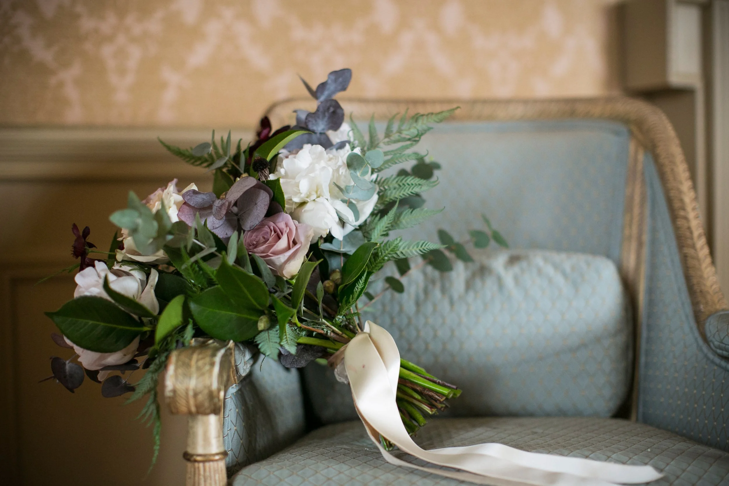 A bouquet of flowers, including white, pale pink roses, and greenery, resting on a vintage light blue cushioned sofa with gold accents in a decorated interior setting.