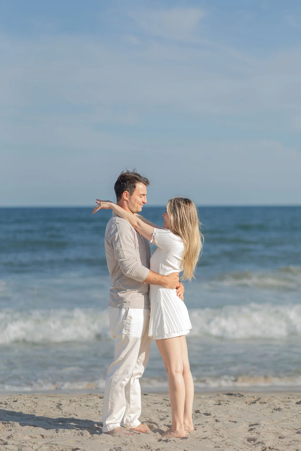 A couple stands on a sandy beach facing each other, holding each other and smiling, with the ocean and blue sky in the background during daytime.