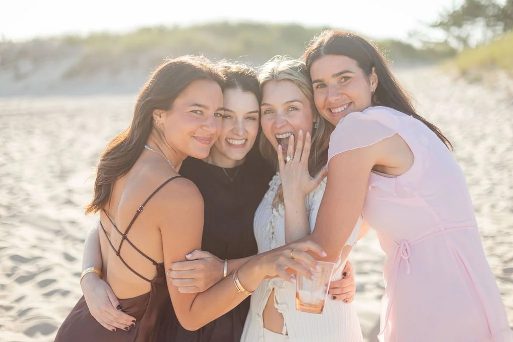 Four women smiling and hugging on a sandy beach with sunlight in the background.