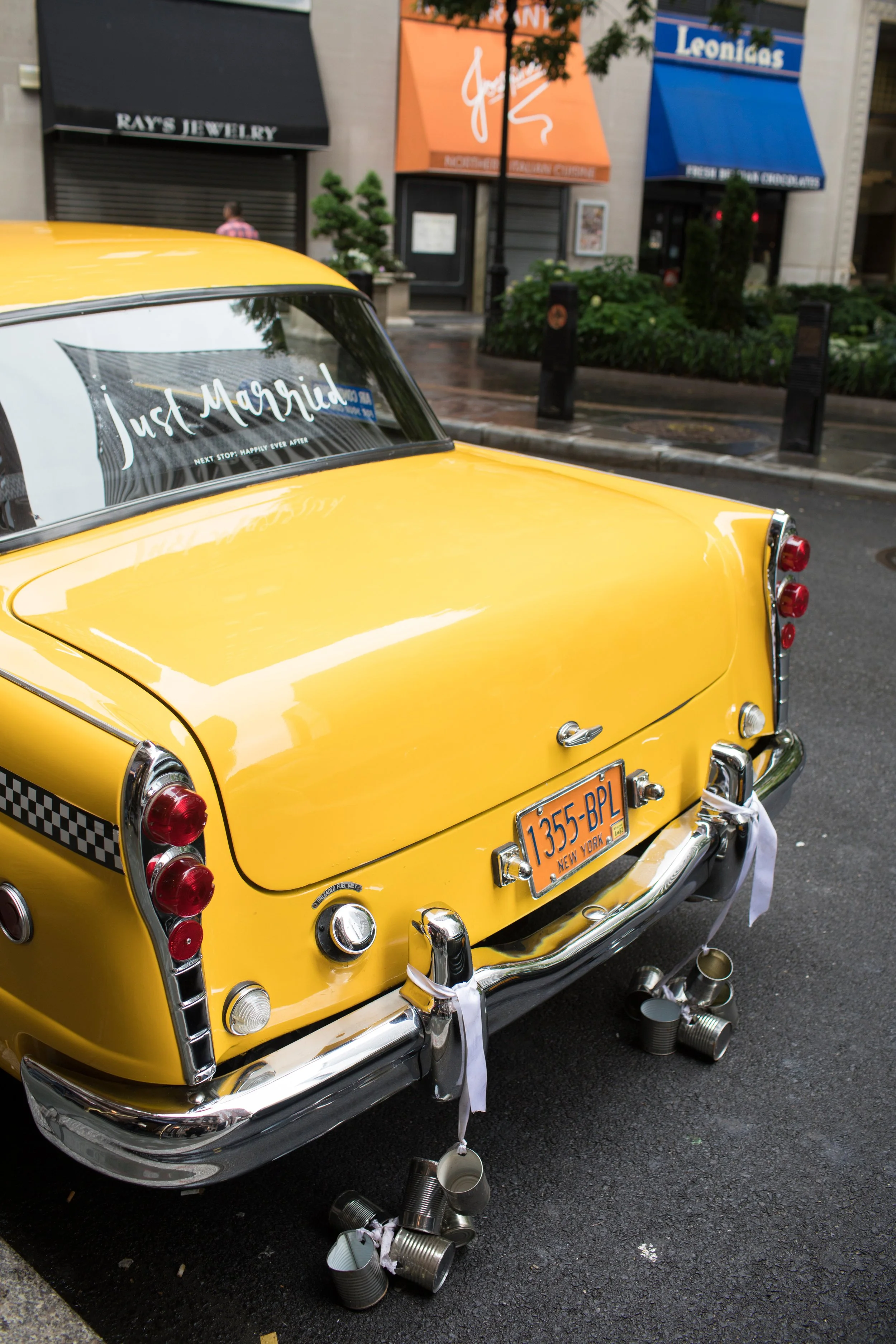 Yellow vintage taxi cab with wedding decorations and a sign saying 'Just Married' in the back window, parked on a city street.