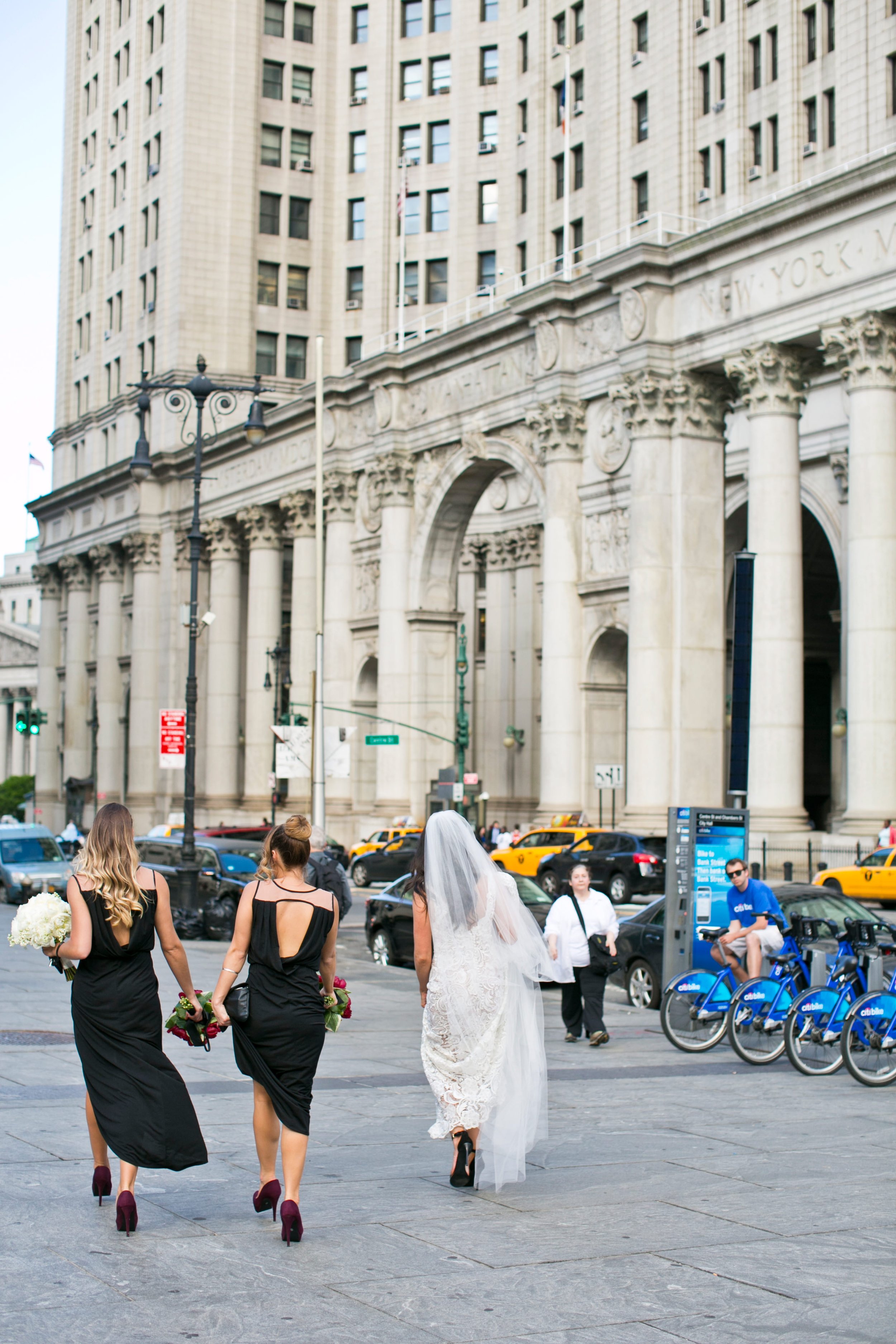 A bride in a white lace wedding dress with a veil and black heels walking with three bridesmaids in black dresses and high heels on a city sidewalk, with a large stone building and taxis in the background.